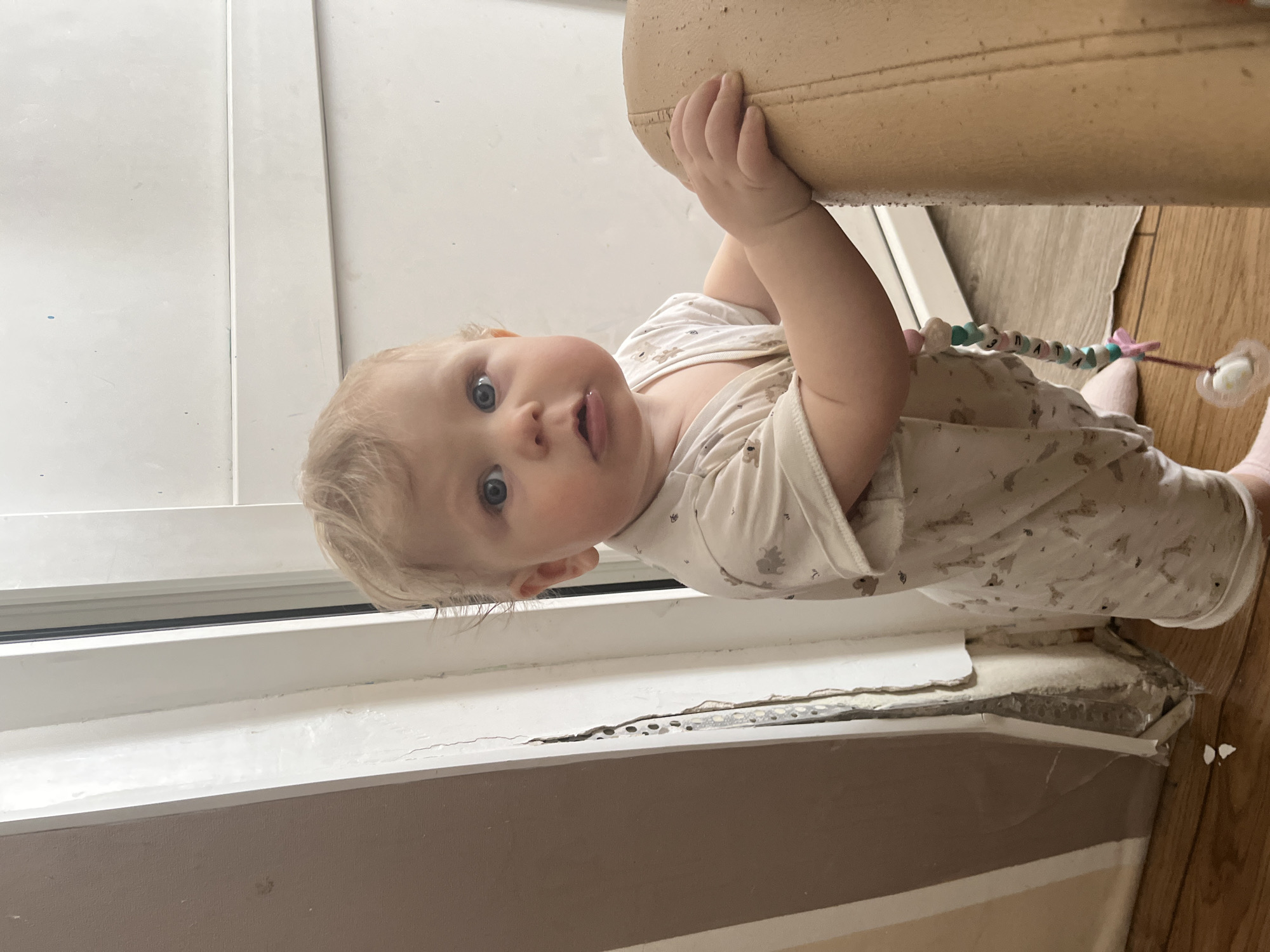 A baby with blond hair and blue eyes, wearing a patterned onesie, is holding onto a cylindrical object. The baby is looking directly at the camera. A pacifier is visible near the baby's leg. The background includes a white wall with visible damage and what appears to be a window frame. The scene is located in an interior space. The location in the city of Oryol-Izumrud, Russia is unknown.