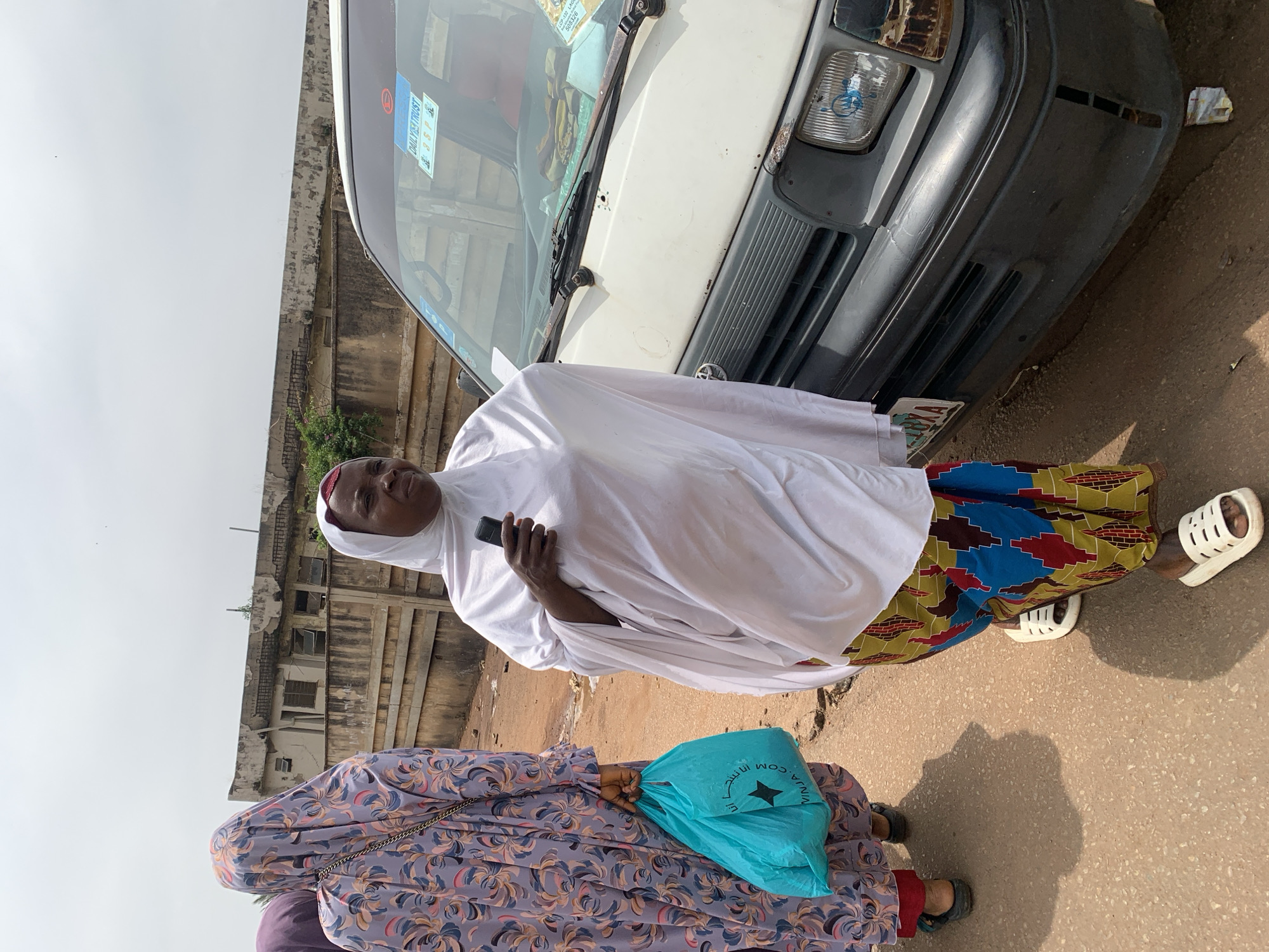 In a street scene in Jos, Nigeria, two women walk alongside a white van. The woman in the foreground, wearing a white hijab and a colorful, patterned skirt, holds a dark cell phone in her right hand. She walks with white slide sandals. To her left, another woman, whose face is obscured by a light purple hijab with a floral pattern, carries a bright turquoise shopping bag in her left hand. She wears black sandals. The van, with a slightly rusted front, has a license plate that appears to read "35 P". Stickers are visible on the windshield, including one that says "DAILY TRUST". The background shows a weathered, light-colored building with rows of windows, and a concrete wall. The ground is a reddish-brown dirt and asphalt mix. The sky is overcast.
