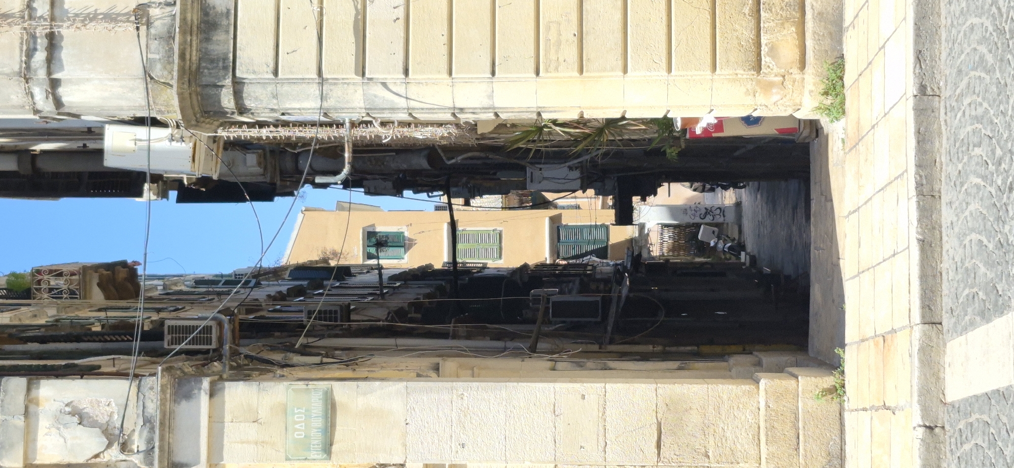 The image depicts a narrow street in Corfu, Greece, with buildings on either side. The street appears to be paved with cobblestones and flagstones. On the left side, there is a building with a sign that reads "ΟΔΟΣ ΕΥΓΕΝΙΟΥ ΒΟΥΛΓΑΡΗ" (Odos Evgeniou Voulgari), indicating the name of the street. The building on the left exhibits signs of age with chipped paint and exposed plaster. Numerous air conditioning units and electrical wires are visible on the facades of the buildings, creating a cluttered appearance. On the right side, a light-colored stone building with horizontal ridging is prominent.

Further down the street, a building with yellow painted walls and green shutters is visible. The alleyway leads into the distance, where a security gate and two parked motorcycles can be seen. The sky above is clear and blue. The scene conveys a sense of dense urban architecture typical of older European cities.