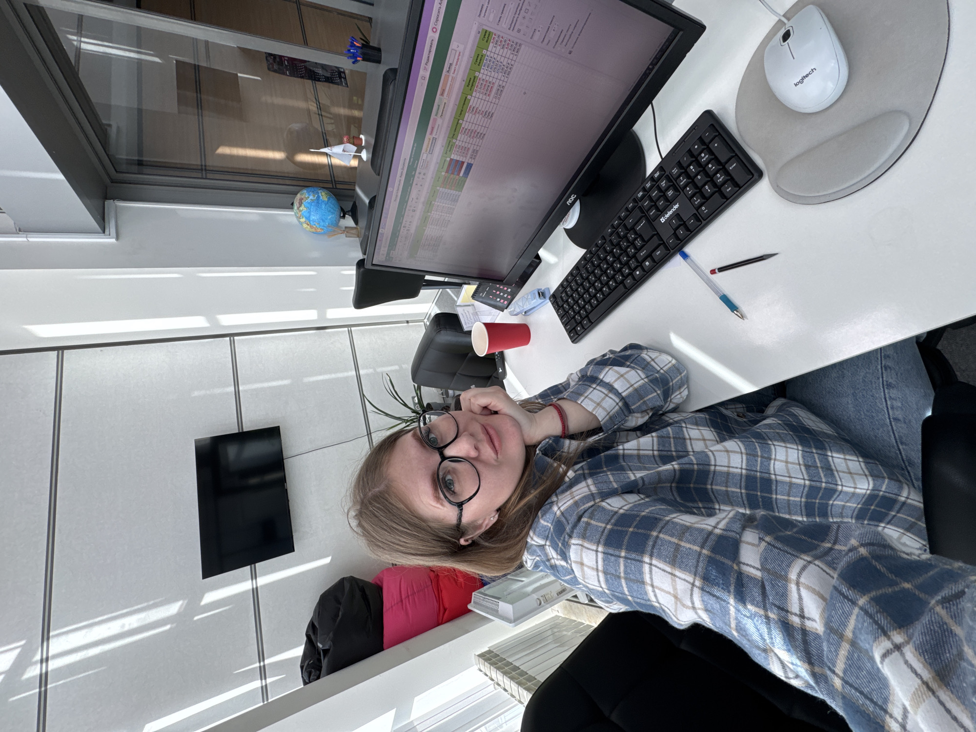 A female individual is depicted in a selfie taken from an office workstation in Perm, Russia. The subject, a young adult with light brown hair, wears black-rimmed glasses, a blue and white plaid shirt, and blue jeans, resting her chin on her right hand while looking at the camera. She is seated at a white desk in front of a black AOC computer monitor displaying a spreadsheet program (likely Microsoft Excel) with text in Russian, including the city name "Пермь". A black Defender keyboard with a Russian layout and a white Logitech mouse on a grey ergonomic mousepad are on the desk, alongside a red disposable cup, a blue stapler, and writing implements. The background features a modern office environment with white walls and horizontal grey trim. Strong diagonal sunlight streaks across the wall from a large window on the right. A black flat-screen television is mounted on the wall, and a stack of three beanbag chairs (one black, two pink) is visible to the left. A white radiator is positioned below the window. Behind the monitor, on a separate dark surface, are a small globe, a miniature white flag, a potted plant, and a pen holder.