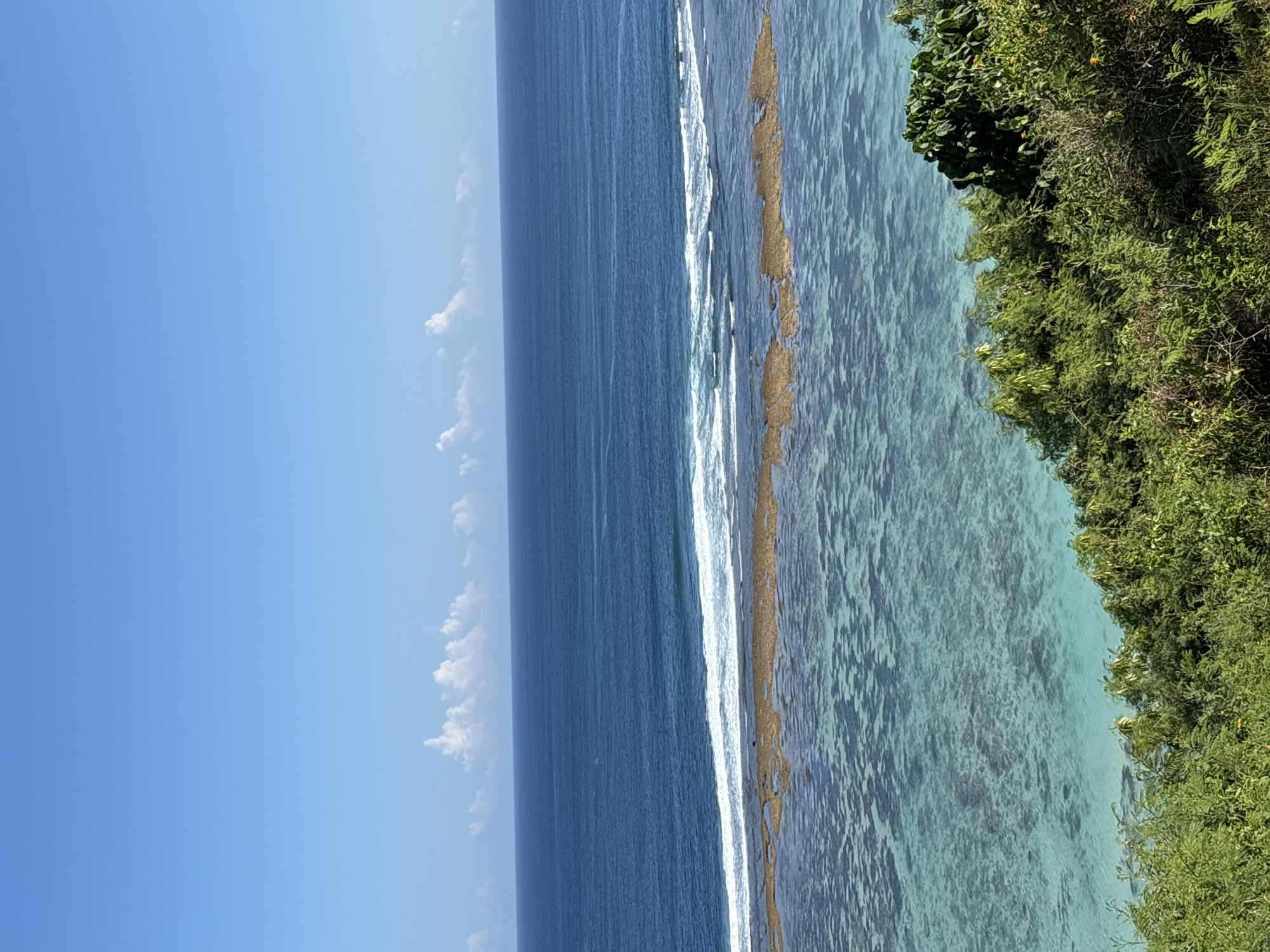 The image depicts a coastal view, likely taken from an elevated position near Benoa, Indonesia, with a bright blue sky occupying the upper left portion of the frame. Thin, white clouds are scattered near the horizon. The ocean transitions from a darker blue further out to a lighter, turquoise color closer to the shore, indicating shallow water. A line of white surf breaks along what appears to be a coral reef or sandy bottom, separating the deeper blue water from the shallower turquoise area. Lush green vegetation, possibly a cliffside or hillside, occupies the right side of the frame. There are no visible people or animals in the scene. The clear visibility suggests a sunny day with good weather conditions.