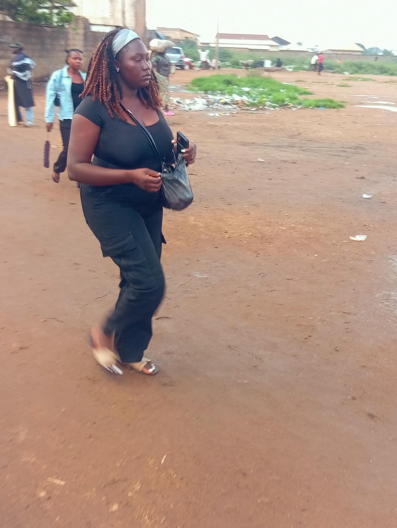 A woman with braided hair, wearing a black t-shirt and black pants, walks across a dusty, dirt path. She has a grey and white headband in her hair and a black purse slung over her shoulder. In her right hand, she holds a cell phone. She is wearing open-toed sandals. Behind her, another woman in a light blue denim jacket and black pants walks with a purple umbrella. Further in the background, other individuals and a parked vehicle are visible. The scene appears to be an outdoor area with scattered debris and vegetation. The lighting suggests it might be daytime, possibly late afternoon. The overall impression is a candid street scene in an outdoor setting, likely in Bukuru, Nigeria.