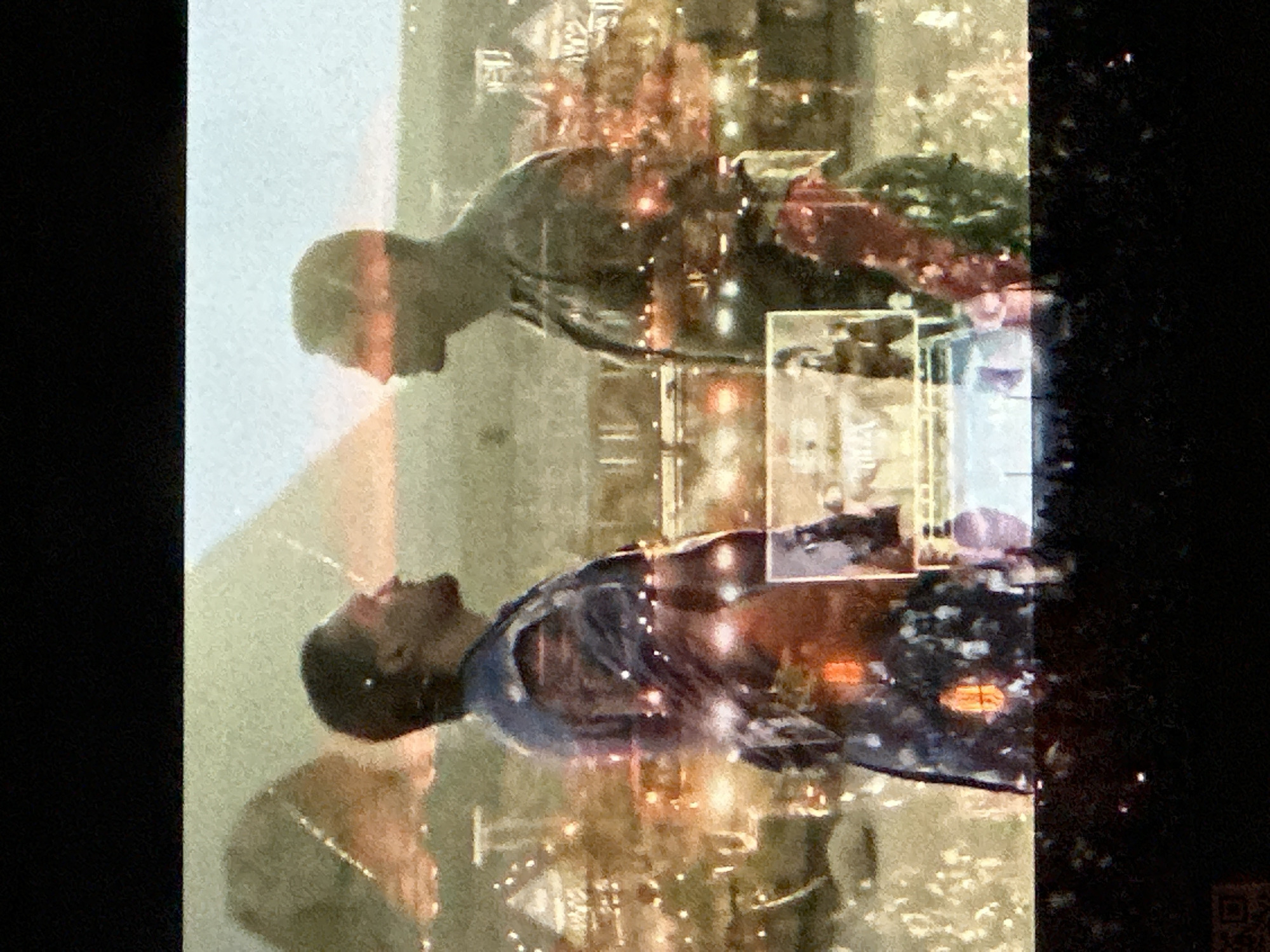 The image appears to be an upside-down projection or reflection capturing a scene within an urban environment, possibly North Kuta, Indonesia. Two individuals are visible: one wearing a blue top, the other in a gray top. The background includes buildings and city lights, suggesting a nighttime setting. A rectangular object is visible, possibly a sign or advertisement. The right side of the image is obscured by darkness. The reflection is blurred and layered, creating a distorted effect.