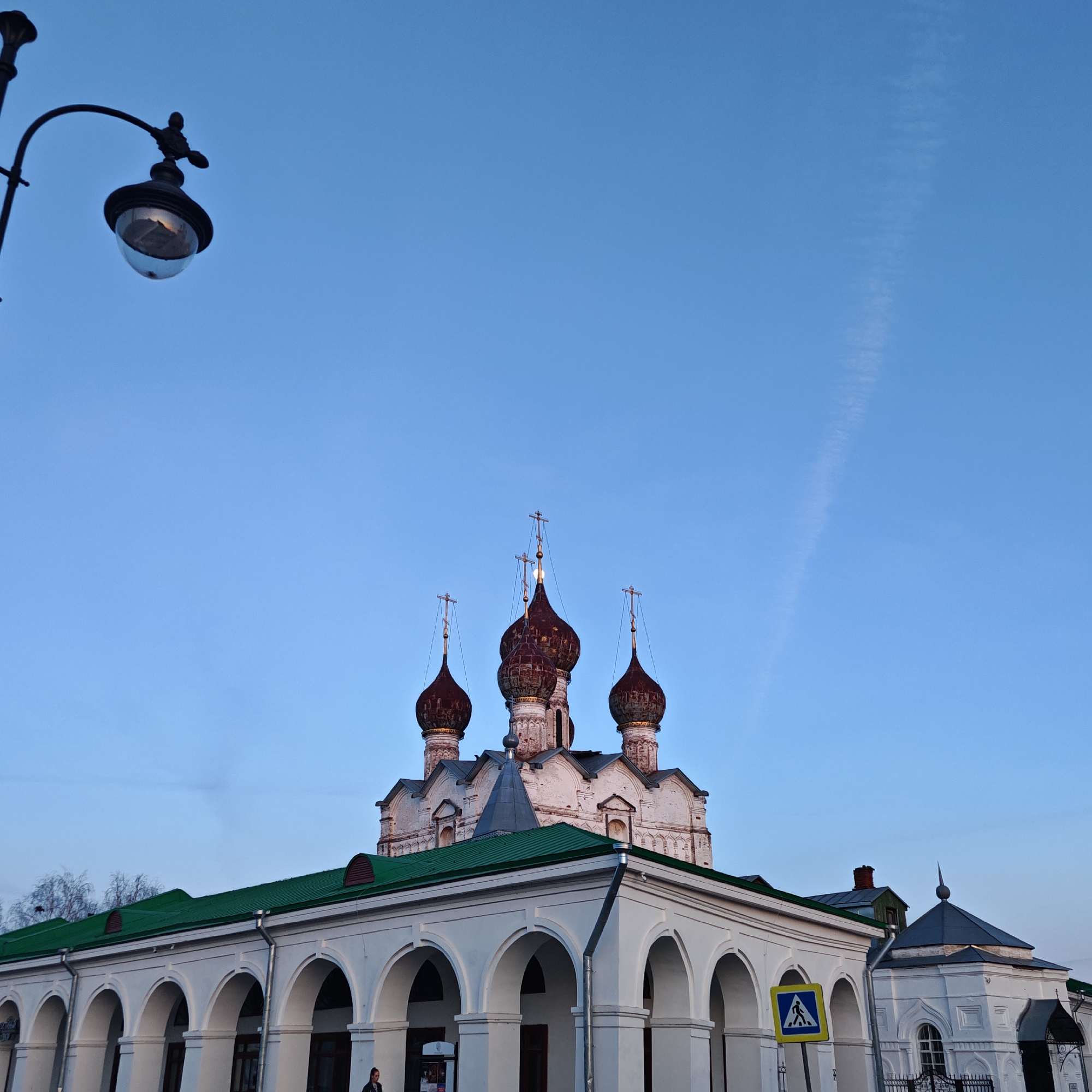 A low-angle shot depicts a white building with arched columns and a green roof in Rostov, Russia, partially obscuring a church with ornate, reddish-brown onion domes topped with crosses. A lamppost is visible in the upper left. The sky is a clear blue with a vapor trail. A pedestrian crossing sign is visible in front of the white building.
