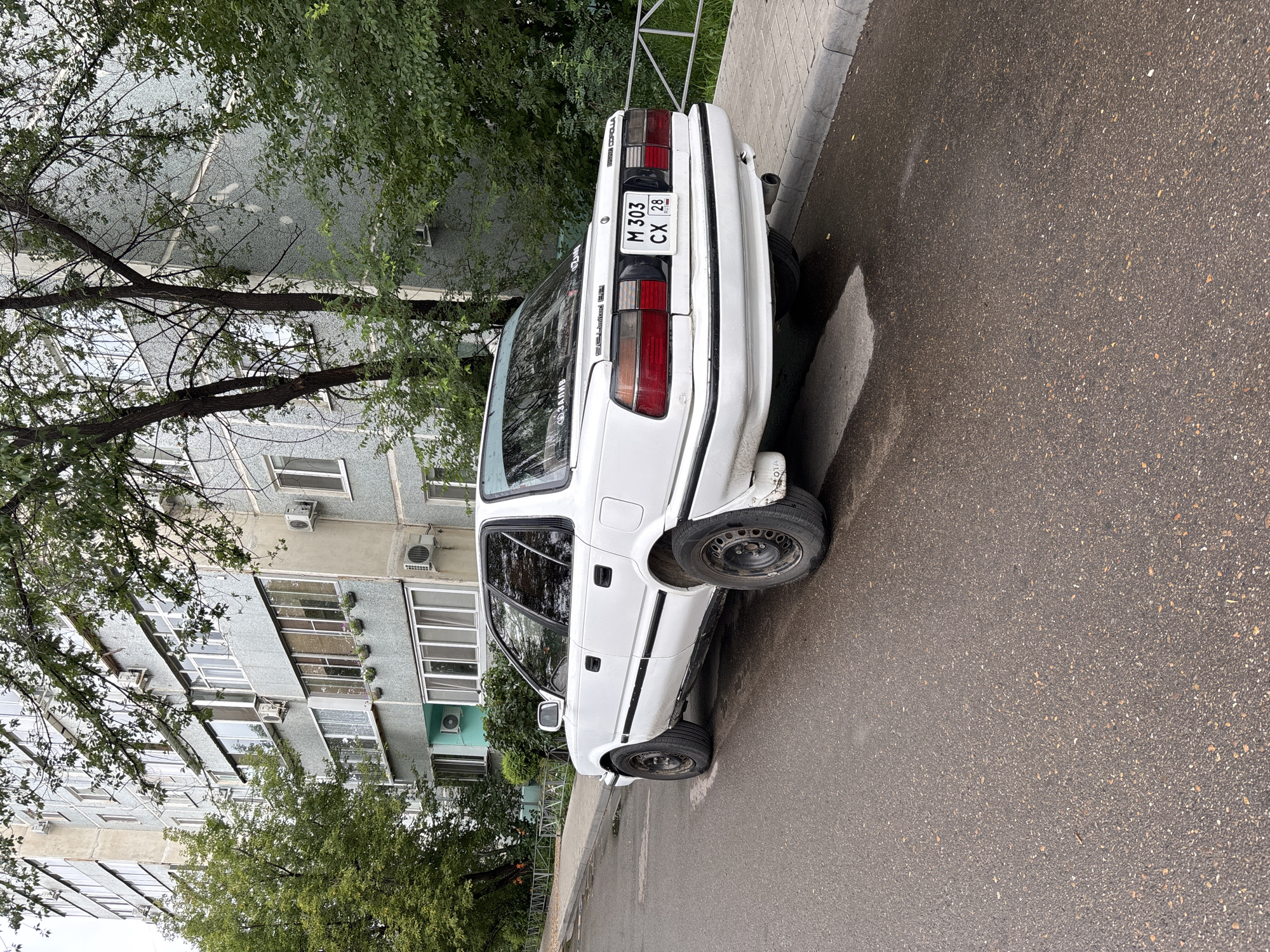 A white Toyota Corolla sedan is parked on the side of a wet asphalt road in front of a grey multi-story apartment building. The car's rear license plate reads "M 303 CX 28", indicating it is registered in Russia. Trees with green foliage grow around the building and partially obscure it. The sky is overcast. A concrete sidewalk with a metal railing runs along the right side of the road, and the car is partially on the curb. The image was taken in Blagoveshchensk, Russia.