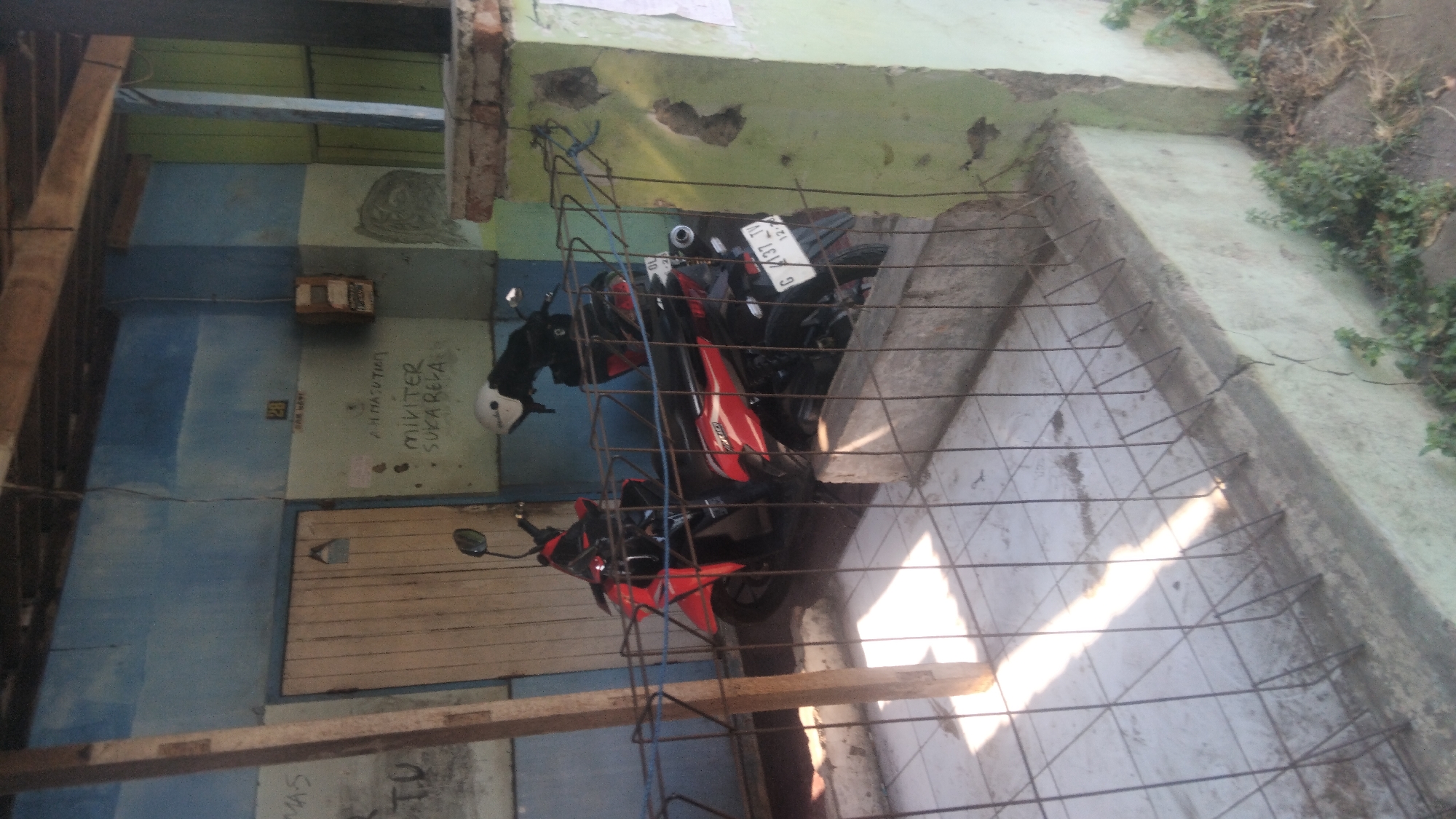 In the city of Proyonanggan Tengah, Indonesia, an outdoor scene depicts a weathered building with a light blue and green facade. A wooden door, slightly ajar, is visible. Two motorcycles, one red and black, the other black, are parked side-by-side in front of the building, partially obscured by a metal fence. The ground is paved with tiles. Graffiti is present on the wall, including the words "AH. HASUTIM MIVITER SUKA RELA" and the number "28" in a small box. An electrical box is mounted on the wall. The scene is illuminated by sunlight casting shadows.