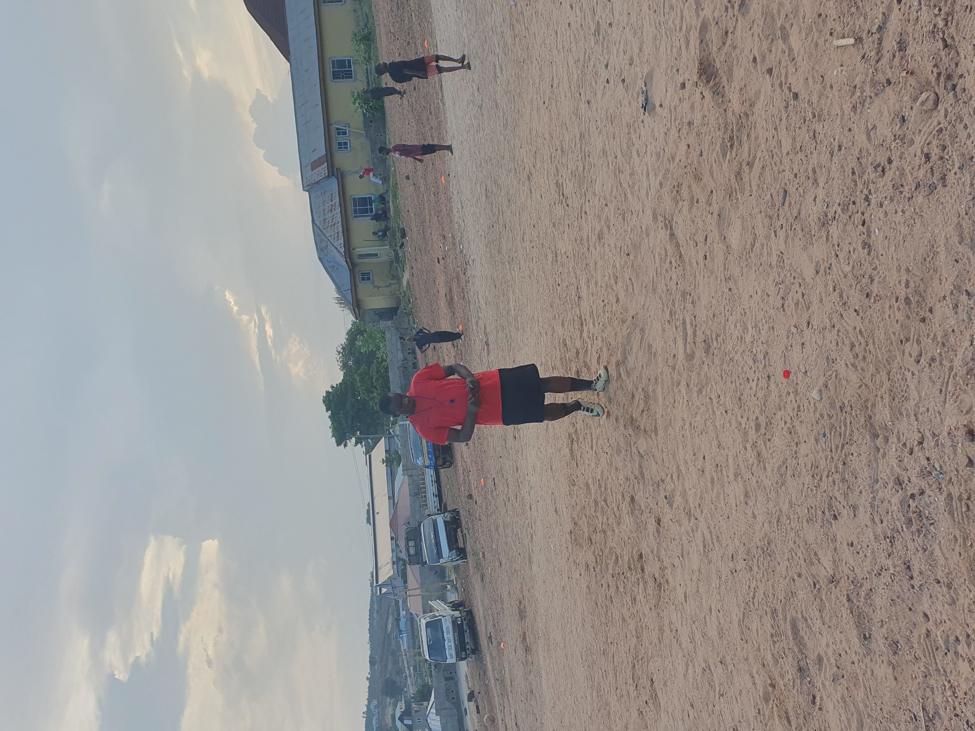 The image shows a sandy field in Lokoja, Nigeria. A man in a red shirt and black shorts stands in the foreground. In the background, several other people are dispersed across the field. A yellow, multi-story building is to the left. There are trucks parked along a road in the background. The sky is overcast.