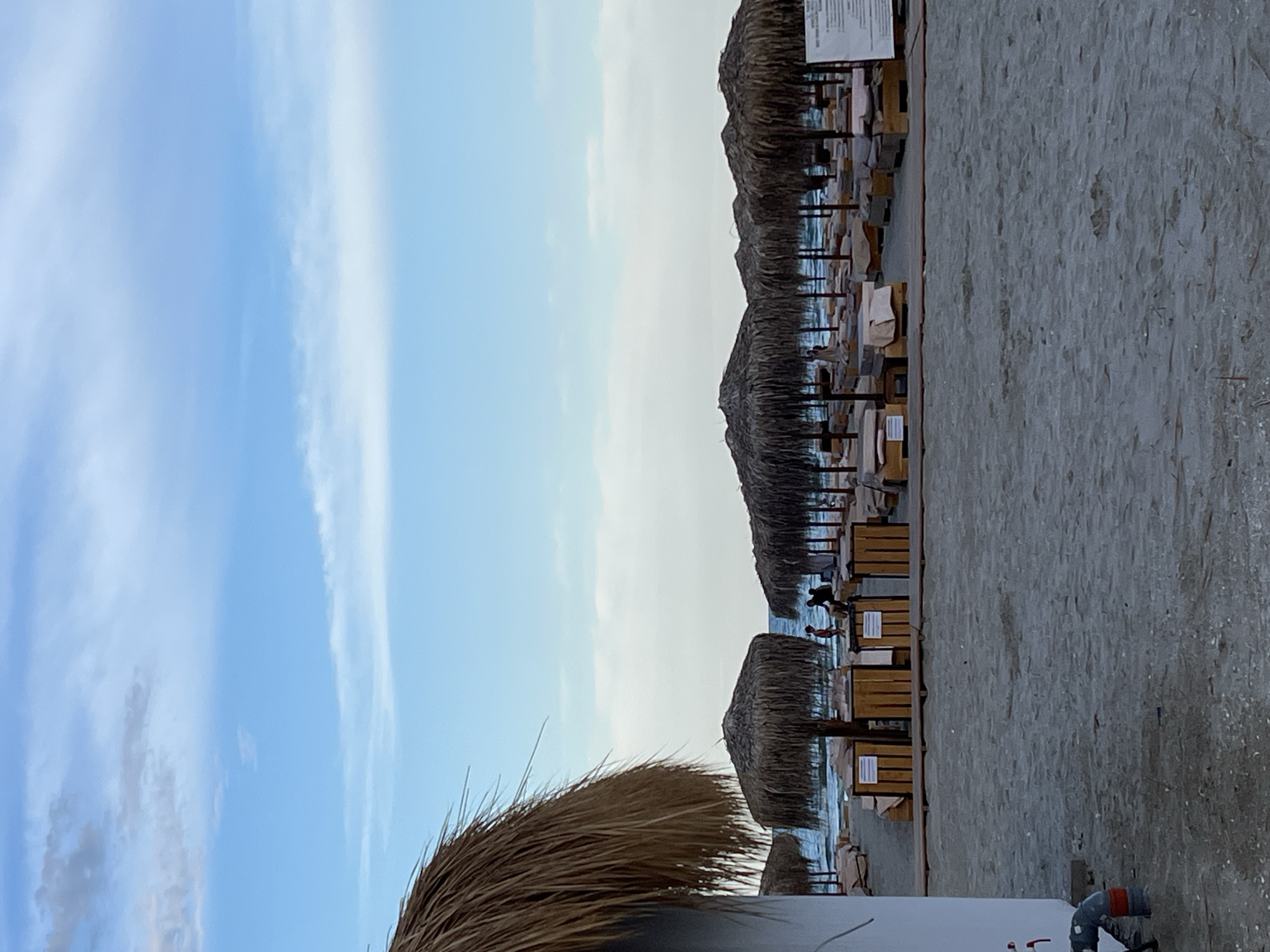 A wide shot captures a beach scene on a cloudy day in Năvodari, Romania. In the foreground, a large portion of a thatched umbrella is visible on the left, partially obscuring the view. The sand stretches out towards the water, where a row of thatched umbrellas and lounge chairs are set up. Several people are visible near the umbrellas and in the water. In the background, the sky is a mix of blue and white clouds.