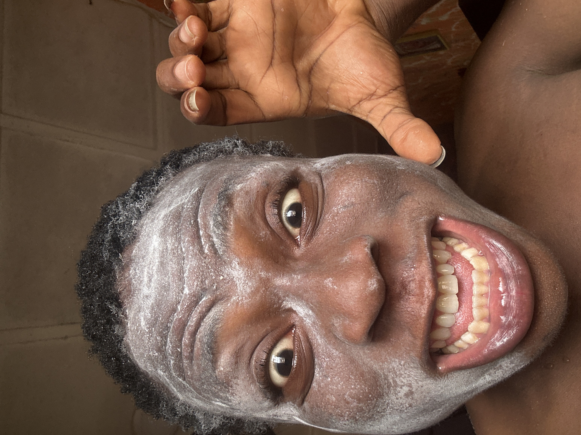 A close-up portrait of a dark-skinned person with a broad smile is presented. Their face appears to be covered in a white, powdery substance. Short, dark hair frames their face. The person's hand, with brown skin and visible veins, is positioned near their head. The background is indistinct, with a mix of brown and white tones. The location is Unknown, Unknown.