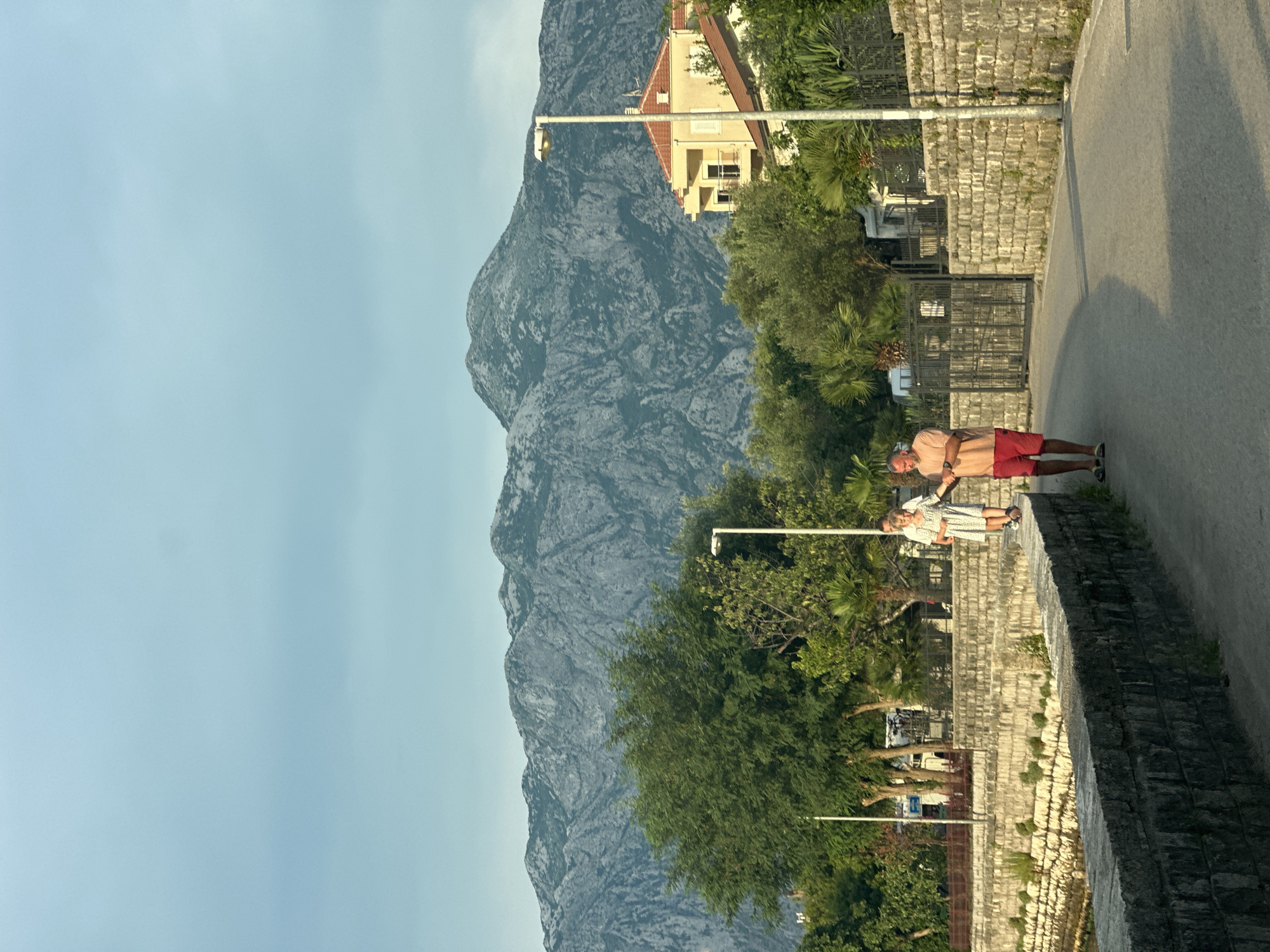 In Prčanj, Montenegro, two people are captured in a casual moment outdoors. A man, wearing a peach-colored t-shirt and red shorts, stands near a stone wall, his arm extended as if interacting with someone nearby. To his right, a woman in a white floral dress is also standing, looking towards him. The scene is set against a backdrop of lush green trees and a prominent, rugged mountain range that dominates the upper portion of the image. The sky is a clear, pale blue, suggesting a pleasant daytime. A street lamp is visible near a building with a red roof. The architecture of the buildings and the stone construction of the walls hint at a Mediterranean or coastal European setting. The overall mood appears relaxed and serene, perhaps during a walk or a break. No visible text is present in the image.