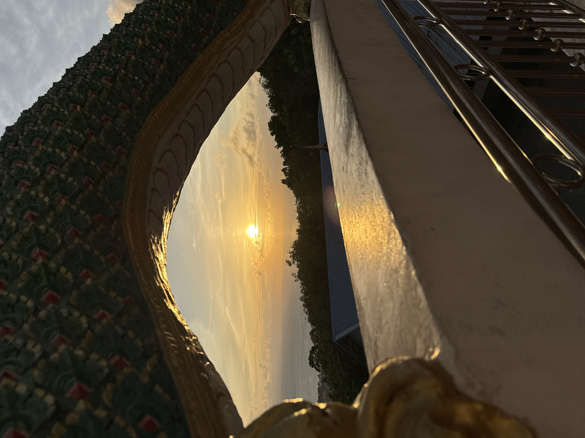 The image shows a sunset over the ocean, viewed through an ornate, serpentine architectural element, possibly part of a temple or a dragon sculpture, at a location in Patong, Thailand. The sun is a bright, glowing orb near the horizon, casting a warm, golden light across the sky and the water. Wispy clouds are scattered across the sky. The ocean stretches out to the horizon, with gentle waves visible. In the foreground, a concrete structure with a metal railing is present, reflecting the sunlight. Lush green foliage covers a hillside in the middle ground, with the roof of a building visible beneath the trees. The architectural element in the foreground is detailed with intricate patterns in green, gold, and red. The overall scene captures a serene and picturesque view of the sunset from an elevated vantage point in Patong.