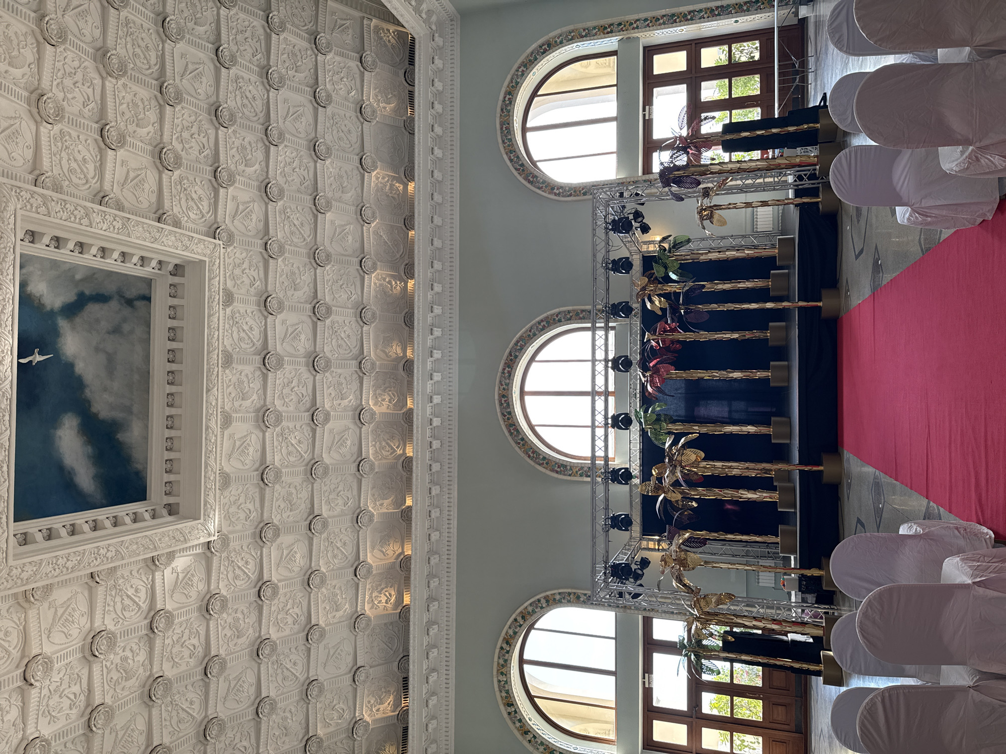 The image depicts a wide-angle view of an indoor event space, possibly a hall or ballroom. The room features an ornate, white, coffered ceiling with intricate detailing and a central rectangular opening painted with a sky and clouds scene, complete with a bird in flight. The walls are a pale mint green, and large arched windows with decorative surrounds are visible.
In the foreground, a vibrant red carpet leads towards a stage. The stage itself is set up with a black backdrop and is adorned with several tall, metallic palm tree decorations in gold, red, and green. Above the stage, a metal truss system is rigged with several spotlights.
To the left and right of the red carpet, rows of white-covered chairs are arranged, suggesting seating for an audience. Wooden doors with glass panes are visible in the background, leading to an outdoor or adjacent area, with greenery visible beyond them.
The scene is well-lit, suggesting it is either daytime with natural light from the windows or the artificial lighting in the hall is active. There are no people visible in the image. The overall impression is of a prepared venue for a formal event, possibly a presentation, performance, or gala. Given the opulent ceiling and the stage setup, it could be a venue such as a historic hall or a banquet room. Without further context, pinpointing the exact location within Sochi, Russia, is not possible solely from the visual information provided.