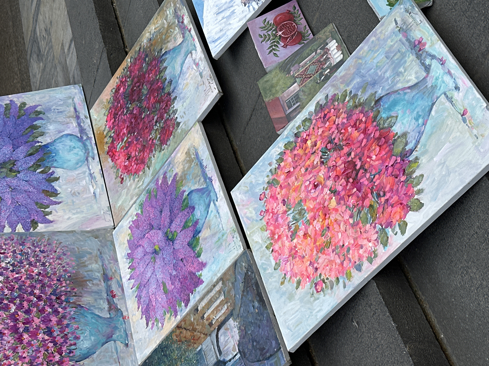 The image shows a collection of small, square oil paintings displayed outdoors, likely for sale. The paintings are resting on a dark, textured surface, possibly concrete steps. The majority of the paintings feature floral still lifes with flowers in vases. The flowers are rendered in thick, impressionistic brushstrokes. Color palettes include shades of purple, pink, and red. There are also paintings depicting pomegranates and a building. The lighting suggests it's daytime, casting soft shadows. The location is likely Tbilisi, Georgia, based on the artistic subject matter and style, common in the area.