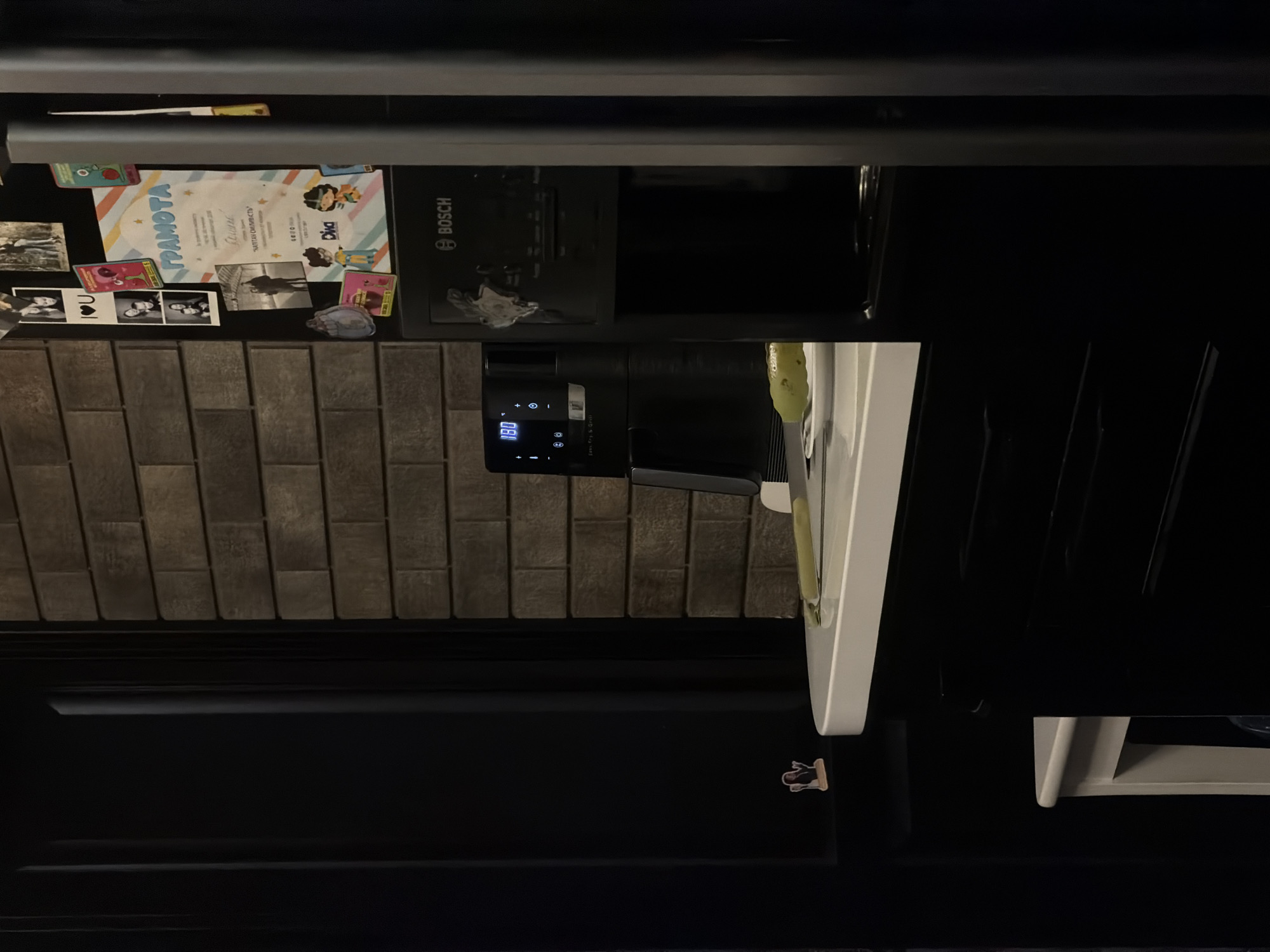 A dimly lit kitchen scene, possibly in Bila Tserkva, Ukraine, features a black refrigerator adorned with photos and a certificate. To the left, a dark cabinet stands against a brick-patterned wall. In the center, a white countertop holds a black air fryer with a digital display showing "180" and a plate with a long, green object, possibly a cucumber. To the right of the air fryer is a black Bosch appliance, likely a coffee maker. The overall ambiance is one of a lived-in domestic space.