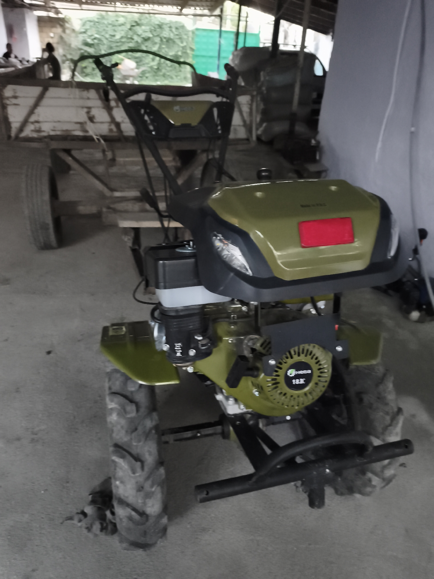 The image shows a green and black rototiller parked indoors. The rototiller has a red reflector on its front. Two people are visible in the background, one sitting and one standing. They appear to be in a warehouse or industrial setting, with sacks of material piled up in the background. The floor is concrete. There is no information in the image to suggest the location is Beynəlmiləl, Azerbaijan.