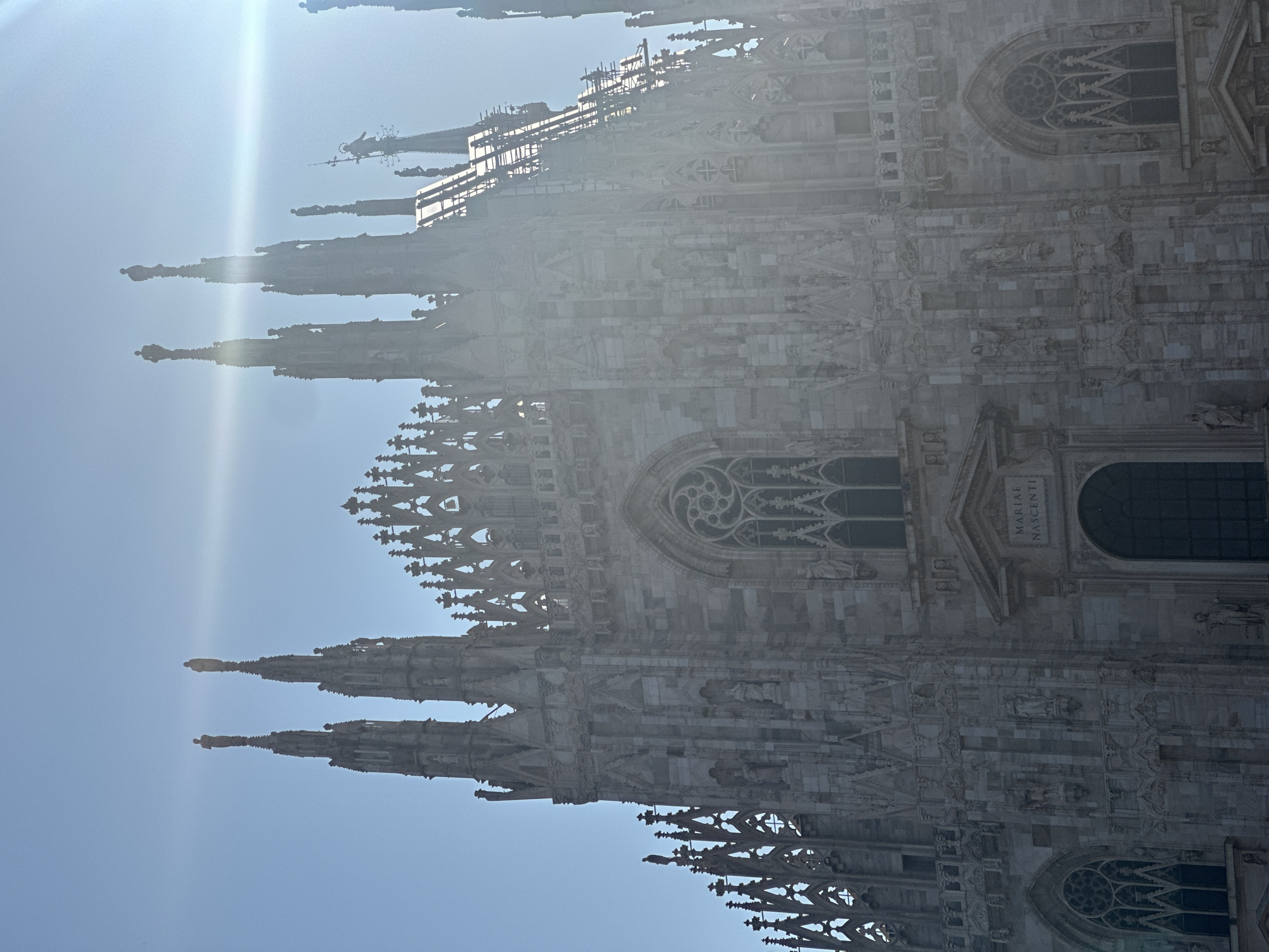 The image shows the Duomo di Milano, also known as Milan Cathedral, in Milan, Italy. The photograph is taken from a low angle, looking up at the facade of the cathedral, which is bathed in bright sunlight, causing some lens flare. The sky is a clear, pale blue.
The Duomo is a magnificent Gothic cathedral characterized by its intricate stone carvings, numerous spires, and detailed facade. The main facade is visible, with several tall, pointed spires topped with statues. The stonework appears to be a light gray marble. In the center of the facade, there is a large arched entrance with a plaque above it inscribed with Latin text, likely referencing the dedication of the cathedral. Flanking this entrance are tall, narrow windows with Gothic tracery. On either side of the central section, more Gothic windows with elaborate designs are visible, along with rows of sculpted figures and decorative architectural elements. The upper portions of the cathedral are partially obscured by the bright sunlight and lens flare, but the characteristic pinnacles and statues of the Gothic style are evident.
There are no people clearly visible in the photograph. The environment is entirely architectural, focusing on the grandeur and detail of the cathedral's exterior. The scene conveys a sense of awe and architectural complexity, highlighting the detailed craftsmanship of the Duomo.