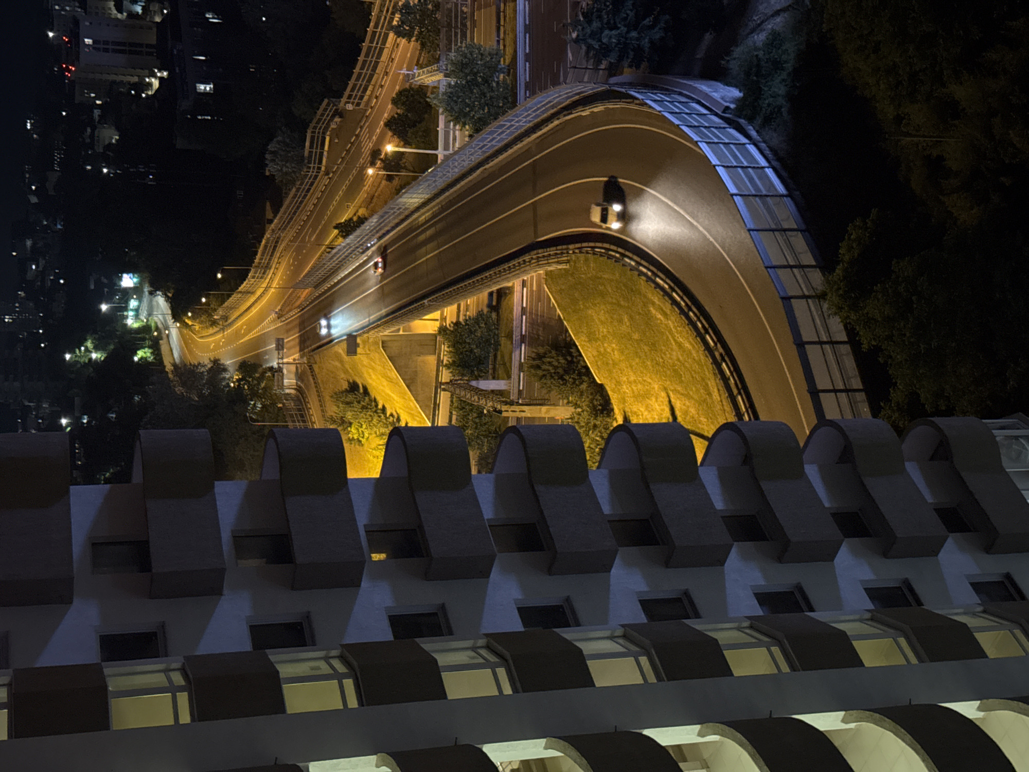 The image captures a nighttime urban scene in Sochi, Russia, focusing on a multi-lane highway curving through a city landscape. A tall, modern apartment building with illuminated windows and balconies dominates the left side of the frame, its facade casting shadows across its balconies. On the right, the highway is illuminated by streetlights and the headlights of moving vehicles. Several cars are visible on the highway, their headlights cutting through the darkness. The highway is bordered by a sound barrier, likely made of glass or translucent material, and surrounded by greenery and scattered buildings in the background. The overall impression is one of a bustling city at night, viewed from an elevated perspective.