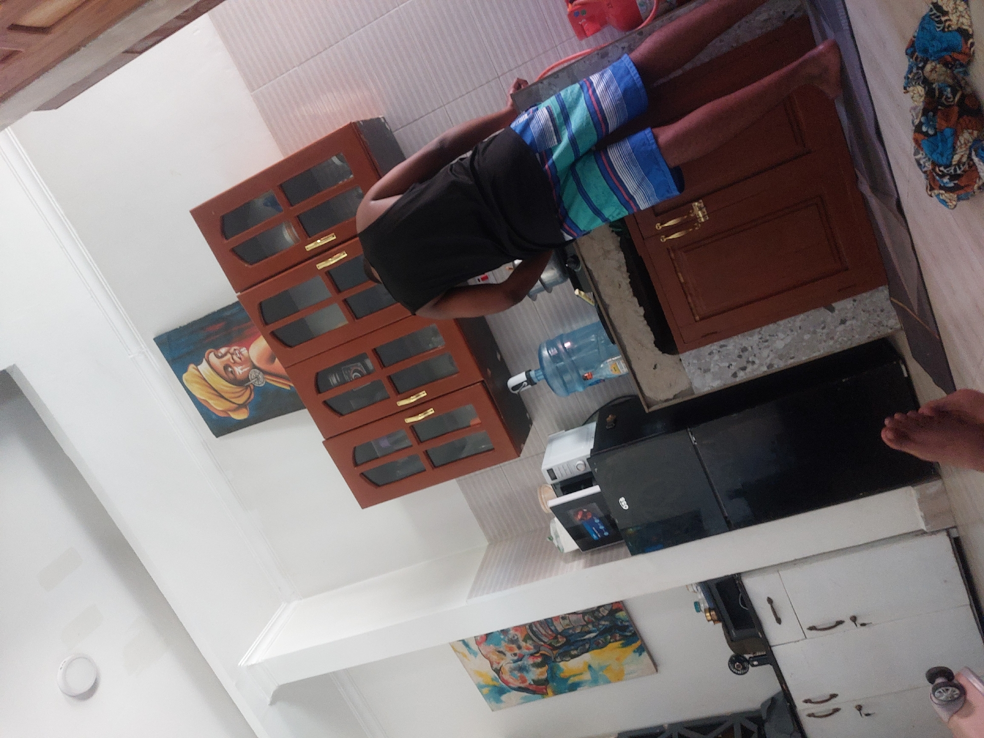 The image shows a kitchen interior. A person wearing a black t-shirt and blue and green patterned shorts stands by the counter. A cabinet with glass doors is mounted above the countertop. On the countertop, there is a water bottle and various kitchen appliances. Below the countertop, there is a black microwave and a white cabinet. Two paintings hang on the wall, one above the upper cabinets and one below the microwave. The specific location in Dar es-Salaam, Tanzania is not discernible from the image.