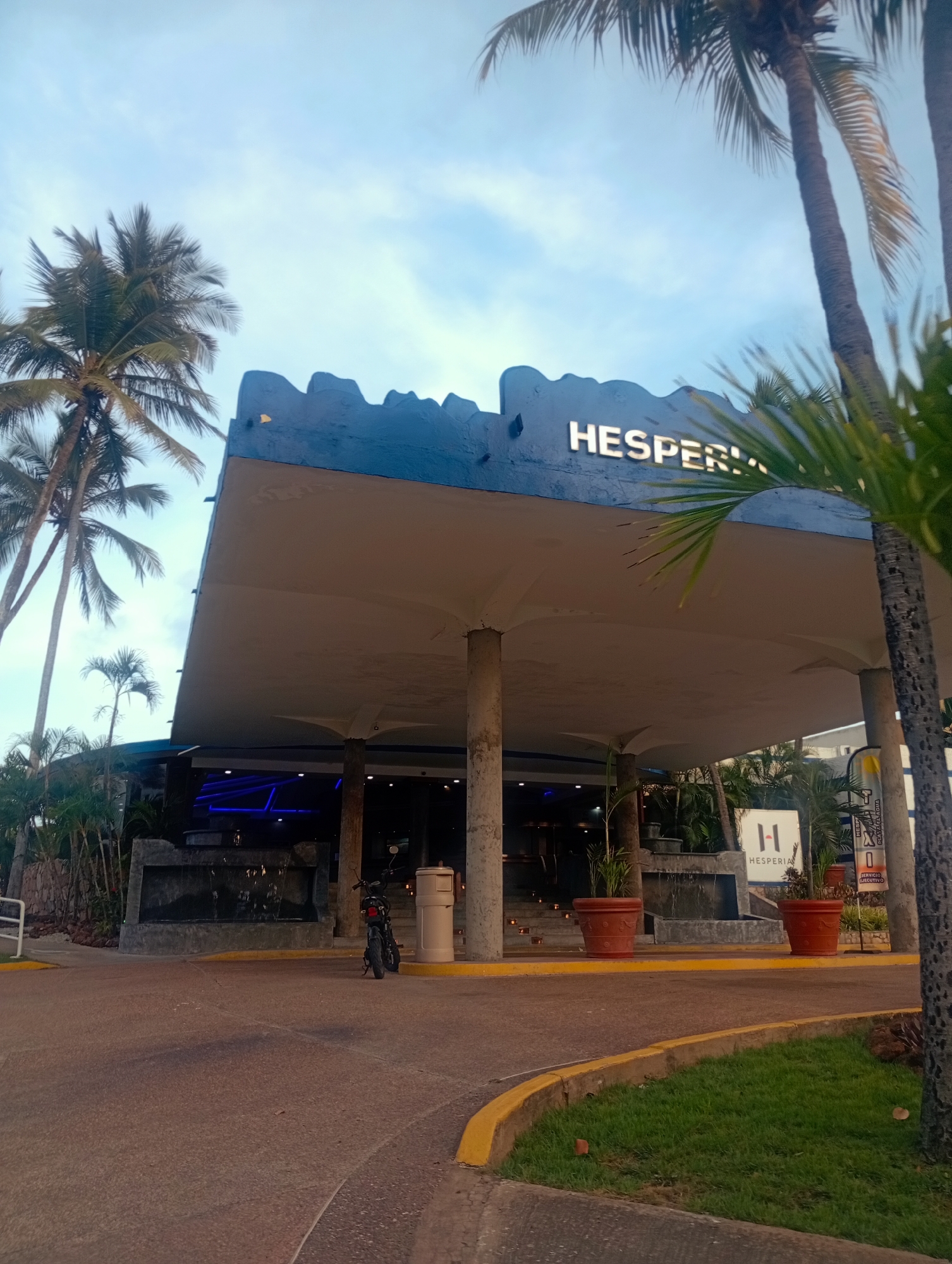 The image depicts the entrance to the Hesperia Playa El Agua hotel in Playa El Agua, Venezuela. The building has a distinctive, modern architectural style with a large, flat overhanging roof supported by cylindrical pillars. The name "HESPERIA" is prominently displayed in illuminated letters on the roof's facade. At ground level, there's a small waterfall feature. A parked motorcycle can be seen near the entrance. Planters with greenery are placed on either side of the entrance. Palm trees are visible in the background. The sky is overcast. A small sign with the hotel's logo is visible near the entrance. The ground is paved.