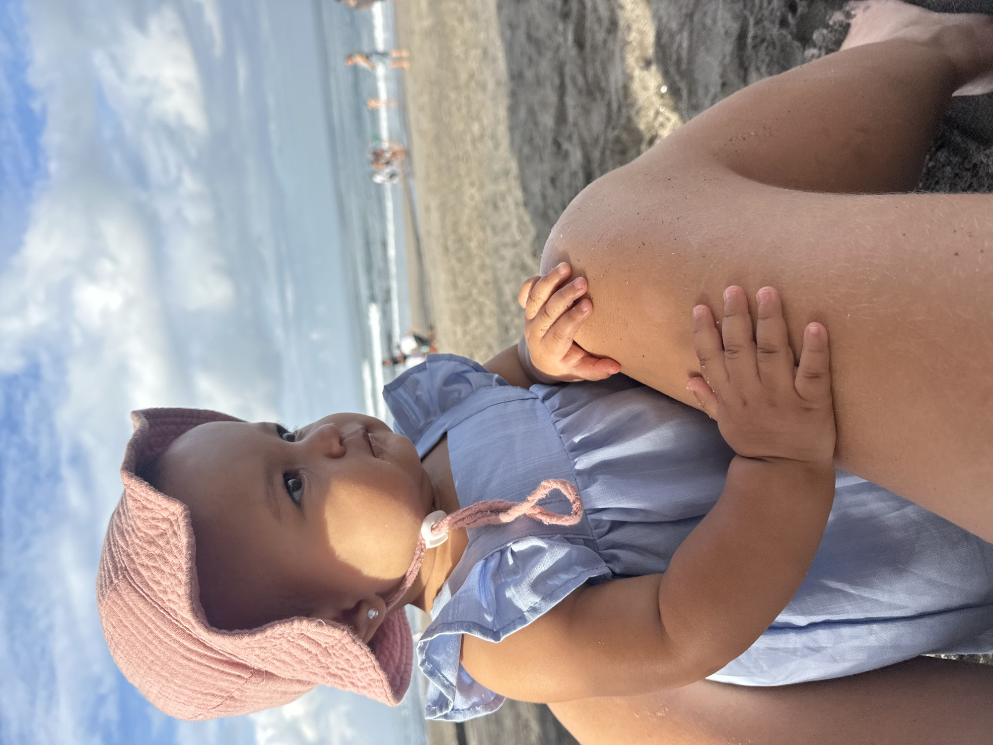 A baby, wearing a pink sun hat and a blue dress, sits on a person's lap on a sandy beach. The baby is looking to the right, with a slight pout. The person's legs are visible, covered in sand. In the background, the ocean meets the sky under a partly cloudy sky. Several people are visible in the water and on the beach in the distance. The location is likely North Kuta, Indonesia.