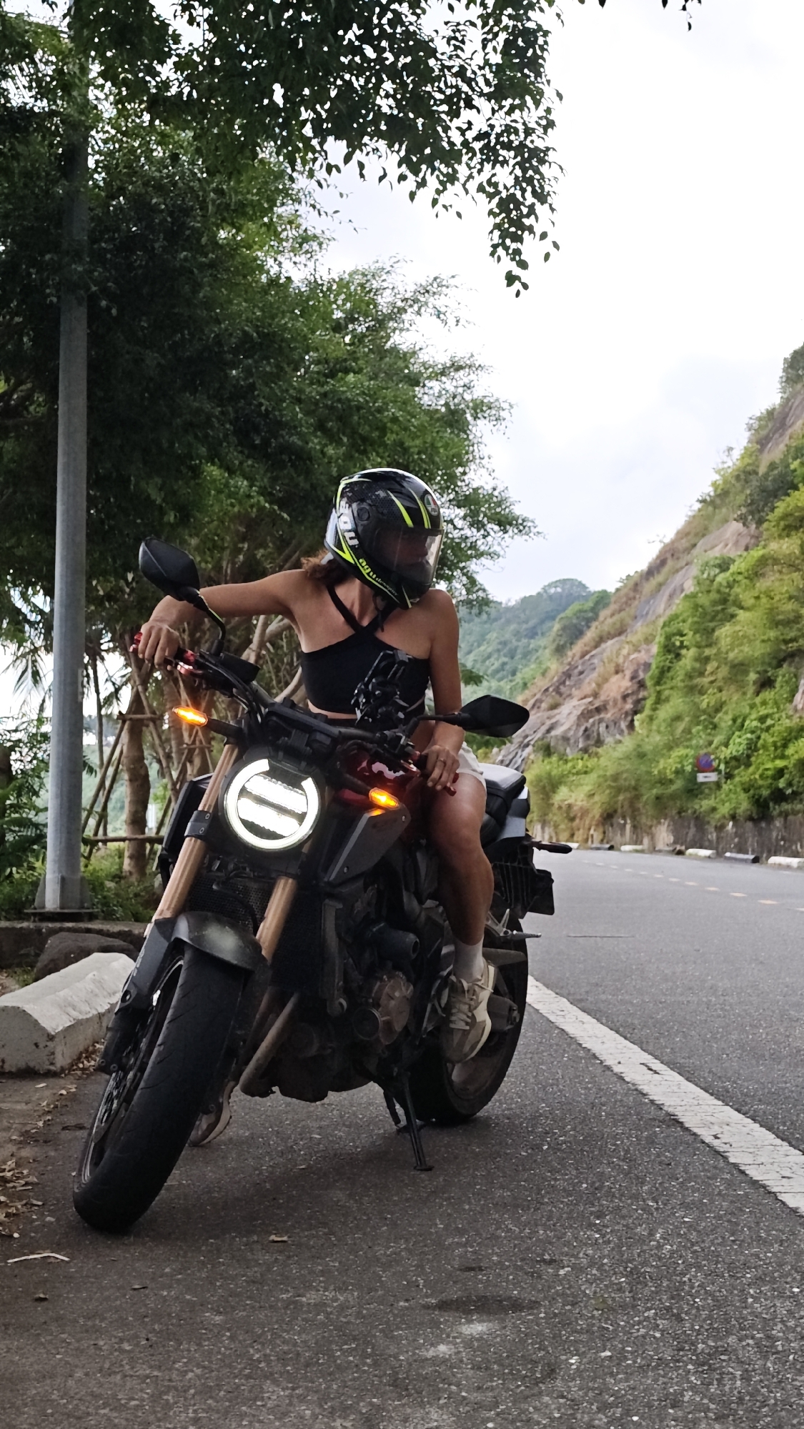A young woman is seated on a motorcycle on the side of a road. She wears a black crop top, light-colored shorts, and sneakers. The woman also wears a black motorcycle helmet with neon yellow accents. The motorcycle has a distinctive round headlight and is parked on a paved road with a white line. To the right, a hillside with vegetation and rock formations is visible. To the left, trees and a lamppost are present. The location appears to be the coastal road leading to Son Tra Peninsula in Đà Nẵng, Vietnam.