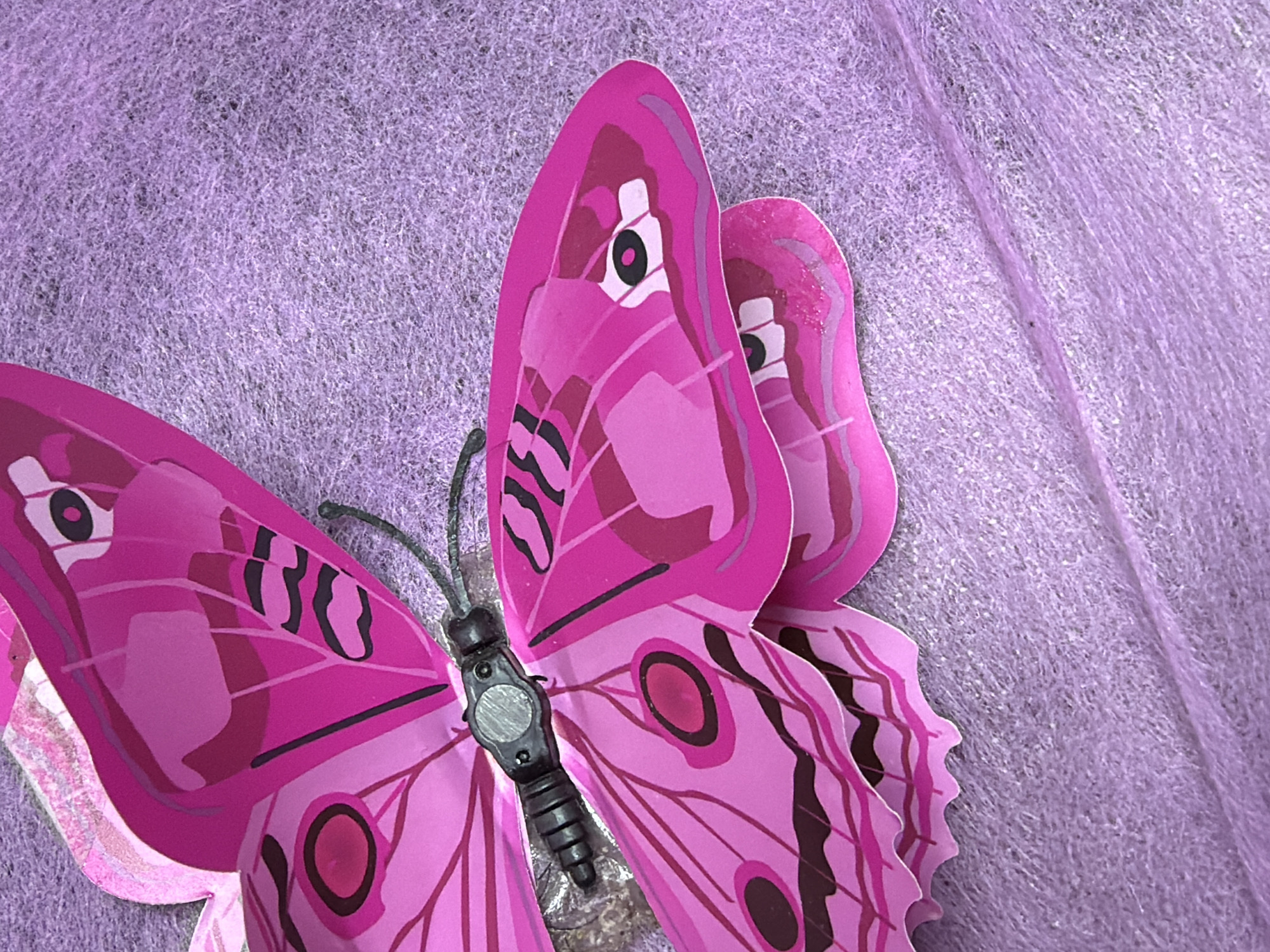 A close-up, overhead shot shows a pink butterfly decoration with black markings on its wings, placed on a textured, light purple surface. The butterfly's body is dark gray with segmented details. The surface appears to be fabric or a similar material with visible fibers. The lighting is even, highlighting the intricate details of the butterfly's wings. There are no people or any other elements that would indicate a specific location within the city of Kyiv, Ukraine, or any other city.