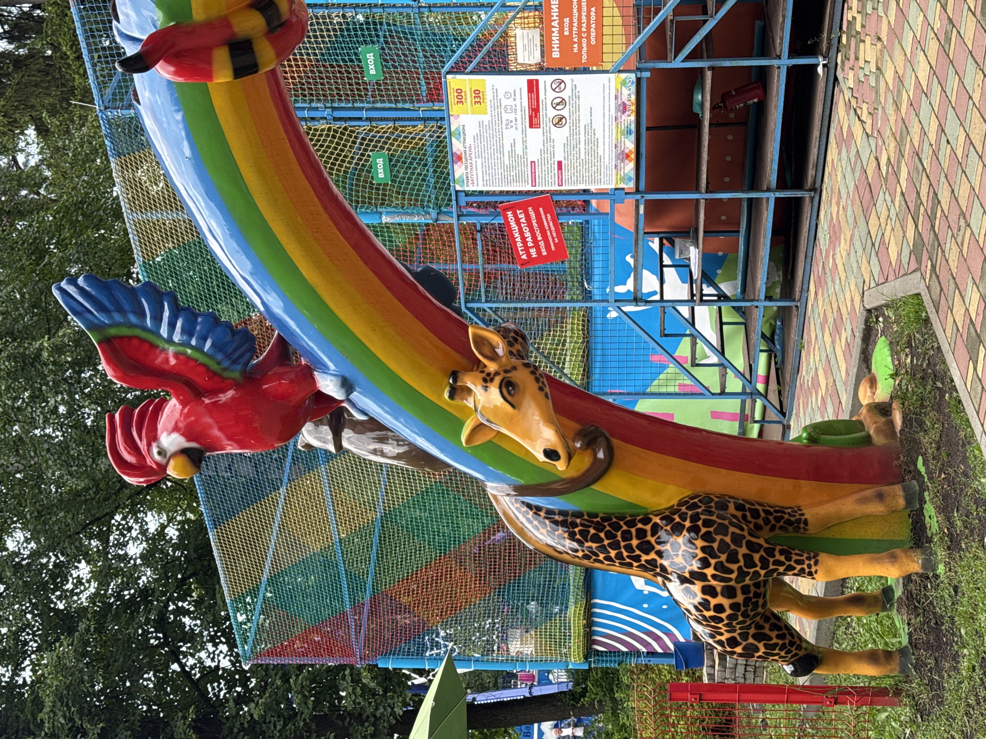 In Izhevsk, Russia, a brightly colored children's inflatable attraction, shaped like a rainbow arch with a red parrot on top and a giraffe supporting the structure, is depicted. A red sign on the attraction reads "ATTRACTION IS NOT WORKING. ENTRY IS PROHIBITED FROM ABOVE." Adjacent to the attraction, a blue metal structure with multiple shelves is visible, containing a fire extinguisher and other items. Informational signs in Russian are posted on the blue structure, detailing rules and prices. The ground is paved with colorful interlocking bricks. In the background, a green umbrella shades an area, and trees with dense foliage create a natural backdrop. The sky is overcast.