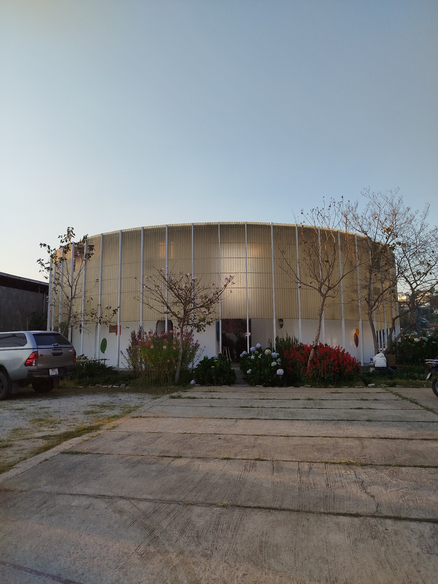 The image shows a circular building, likely a coffee shop, in Da Lat, Vietnam. The building is made of corrugated metal with white vertical supports. The base of the building is white with flowering plants in front. A silver pickup truck is parked on the left side of the image. A concrete parking area is in the foreground. Trees are planted around the building. The sky is clear and light blue. A motorcycle is parked on the right side of the image. Two people are sitting on the ground on the right side of the building.