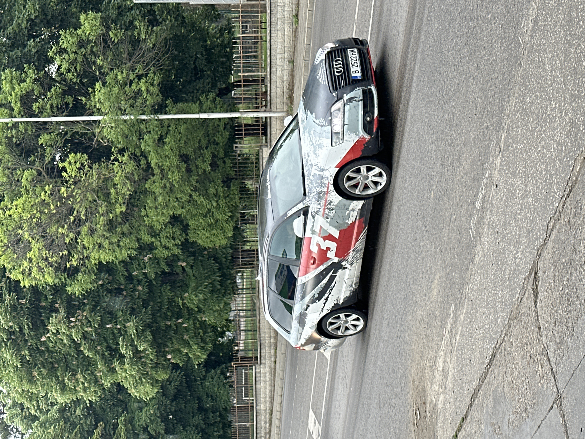 An Audi A6 Avant with license plate B 2522 HM is parked on a street in Veliko Tarnovo, Bulgaria. The car has a unique graphic design with red and white patterns, a British flag motif, and the number 37 prominently displayed. Lush green trees line the street on one side. The road surface is gray asphalt with visible cracks and repair marks. A gray concrete wall runs parallel to the road, behind which a black metal fence is visible. The image appears to be taken from an elevated position, such as a balcony or higher floor.