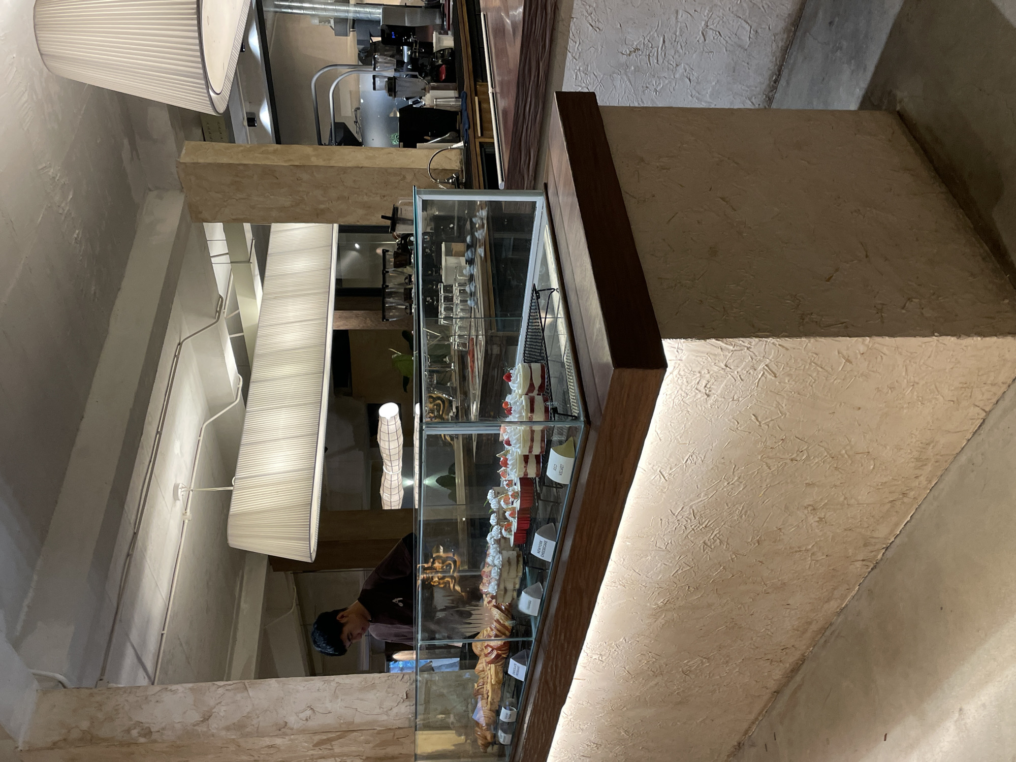A man wearing a brown shirt is seen behind a display case filled with desserts at a cafe. The cafe has a modern industrial design with exposed concrete ceilings and walls. The display case is made of glass and wood, and it showcases various pastries and cheesecakes. The desserts include croissants, red velvet cheesecakes, and other pastries topped with cream and strawberries. The cafe is well-lit with overhead pendant lamps. The overall atmosphere suggests a chic and trendy establishment. The cafe is located in Da Lat, Vietnam, a city known for its French colonial architecture and lush landscapes.