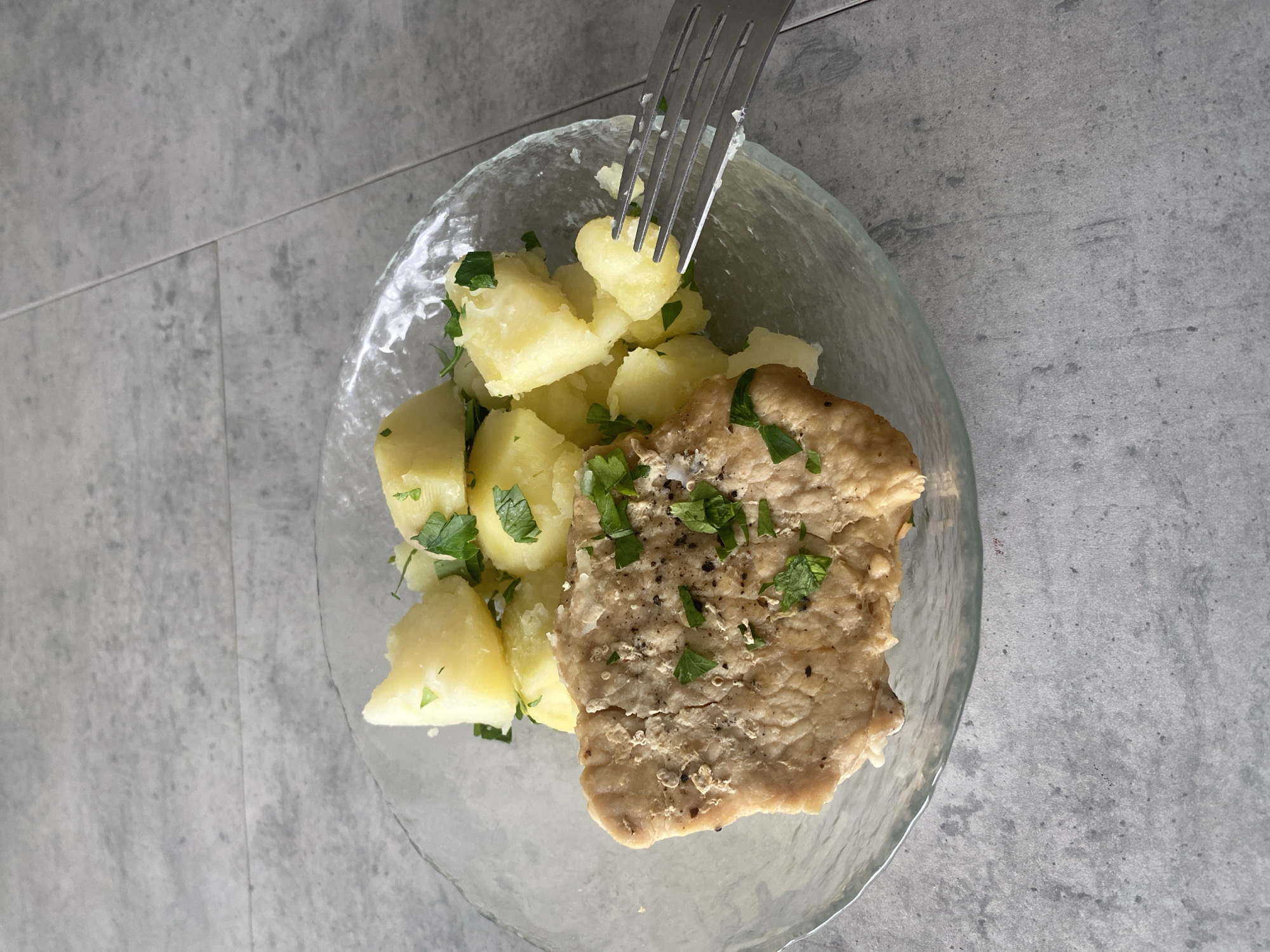 A clear glass plate on a grey, tiled surface holds a meal of boiled potatoes mixed with green herbs, and a single, tan-colored cutlet topped with the same herbs and black pepper. A silver fork is piercing one of the potatoes. The location is not discernible from the image.