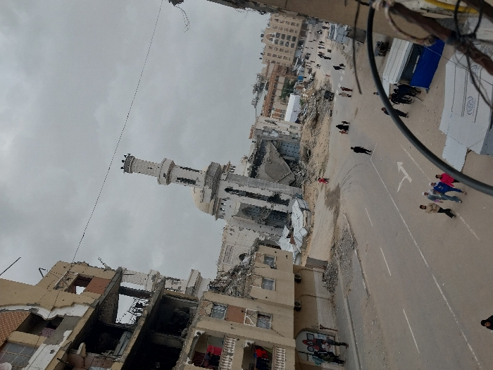 The image depicts an extensively damaged urban street scene in Khan Yunis, Palestinian Territory, under an overcast sky. In the center, the Great Mosque of Khan Yunis is visible, its predominantly white structure and tall minaret standing amidst significant destruction; the main mosque building shows collapsed sections and heavy rubble at its base. To the left, a multi-story building is severely compromised, with large portions of its facade missing, revealing interior rooms and exposed rebar. Scattered debris, rubble, and dirt cover unpaved areas and the sides of a paved road that traverses the foreground and mid-ground. This road features white lane markings and a right-turn arrow. Approximately fifteen to twenty individuals, identified as civilians including adults and children, are walking along the road and its unpaved shoulders in various directions. On the right side of the road, a blue temporary shelter is visible, along with other smaller, makeshift structures constructed from corrugated metal near the damaged buildings. Overhead utility wires span the upper portion of the frame.
