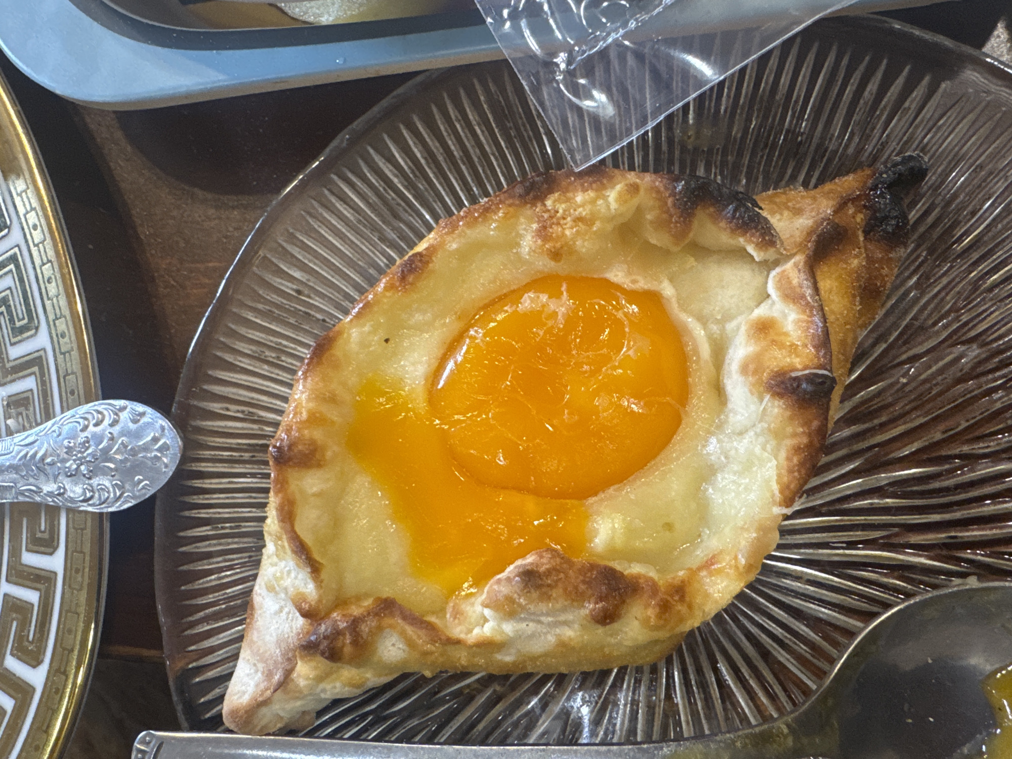 A close-up shot depicts an adjarian khachapuri, a traditional Georgian bread dish, on a clear glass plate with radiating lines. The khachapuri is boat-shaped with a golden-brown crust. The center is filled with a melted cheese and a raw egg yolk. Part of a decorative silver spoon is visible on the left, and a serving spoon with remnants of yolk or cheese is on the bottom right. The location appears to be Yerevan, Armenia, based on the prevalence of the dish in Georgian cuisine and the likelihood of finding it in neighboring countries. The overall scene is a presentation of the dish ready to be eaten, likely in a restaurant or café setting.