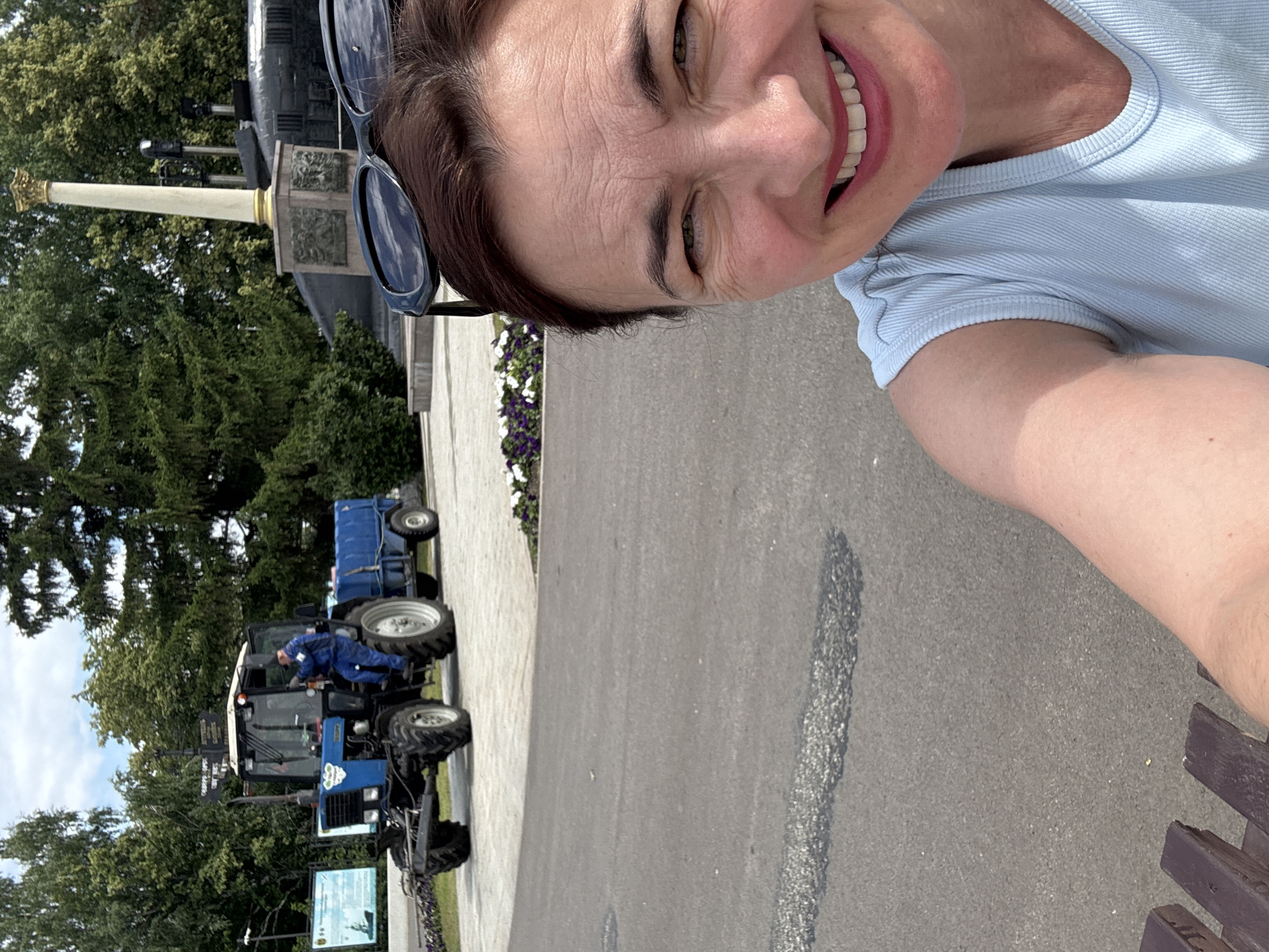 A woman with brown hair and sunglasses is taking a selfie outdoors, smiling at the camera. She is wearing a light blue tank top. Behind her, a blue tractor with a trailer is parked on a paved area. A person in a blue uniform is sitting in the driver's seat of the tractor. The background includes trees, a monument with a column, and a dark structure that appears to be a submarine. The scene suggests a public park or memorial area, potentially in the city of Ufa, Russia, given the presence of a monument and the overall environment. The woman's arm and shoulder are visible in the foreground, and a portion of a wooden bench is also visible.