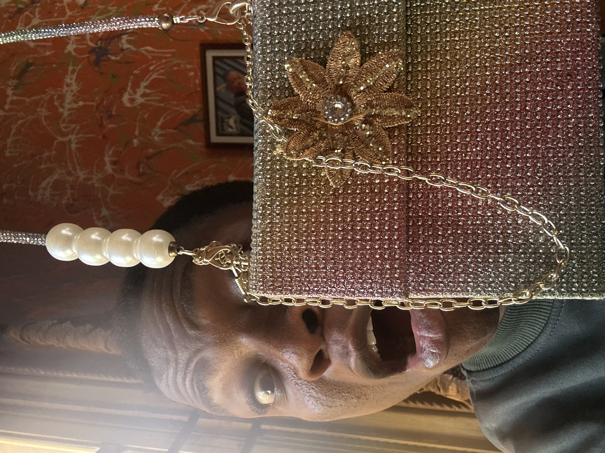 A close-up, portrait-oriented shot features a person with dark skin, whose face and upper torso are partially visible, looking upwards with wide eyes and a slightly open mouth, revealing their tongue. The upper portion of the frame is dominated by a highly embellished, sparkling clutch bag, adorned with small, shimmering gold and silver beads or rhinestones. A decorative golden floral appliqué, featuring small beads and a central clear stone, is prominent on the bag, along with a thin gold chain strap. Another bag strap, made of lustrous off-white pearls and a silver-toned, sparkly chain, is visible near the person's ear. The person wears a grey or dark green ribbed sweater. The background consists of an orange wall with a white, abstract, textured pattern, and a small framed picture containing a blurred image of a person. The indoor setting features diffused lighting, emphasizing the reflective surfaces of the bag and the person's skin. No text is visible.