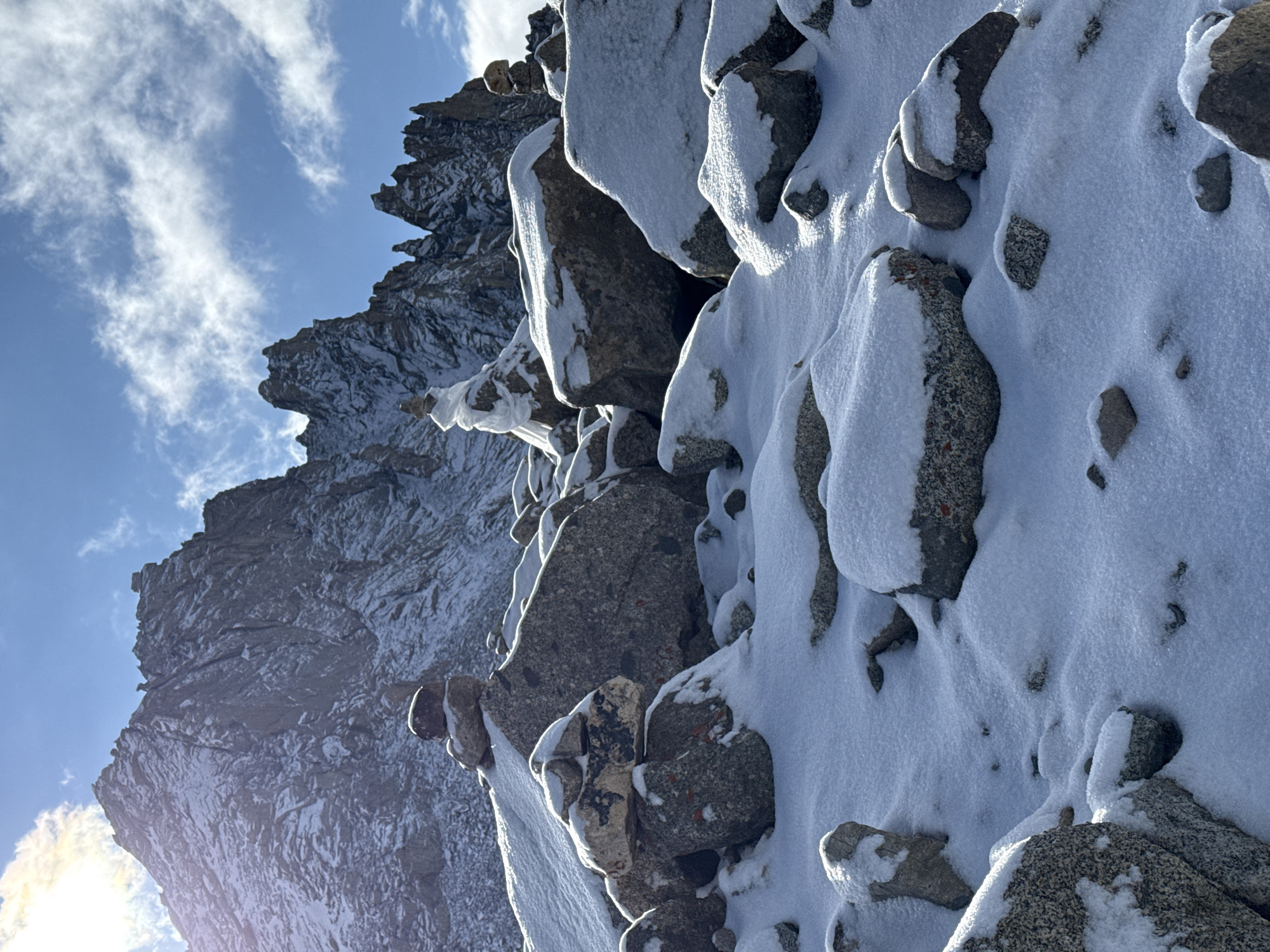 The image displays a rugged, snow-covered mountain landscape under a bright, partly cloudy sky. In the foreground and midground, a steep slope of large, dark grey boulders and rocky outcrops is heavily dusted with fresh, white snow, particularly on their upper surfaces and crevices. The bright sunlight illuminates these snow-covered rocks, casting distinct shadows and highlighting their textures.
To the left of the frame, the scene opens up to a vast expanse of blue sky dotted with scattered, wispy white clouds. Below the sky, in the background, a series of dark, jagged mountain peaks and ridges rise dramatically, also showing patches of snow in their shaded areas. The overall composition suggests a vertical view up a mountain face or across a steep slope.
No human activity, structures, or identifiable objects other than natural elements are present. The time of day appears to be bright daytime, with a strong sun glare visible in the bottom-left corner, indicating the sun is low in the sky, possibly during morning or late afternoon. The weather is clear and cold, as evidenced by the snow and crisp lighting. The scene evokes a sense of remote wilderness, grandeur, and natural beauty in a high-altitude environment. There is no visible text in the image. The location context provided is Parga town, China.