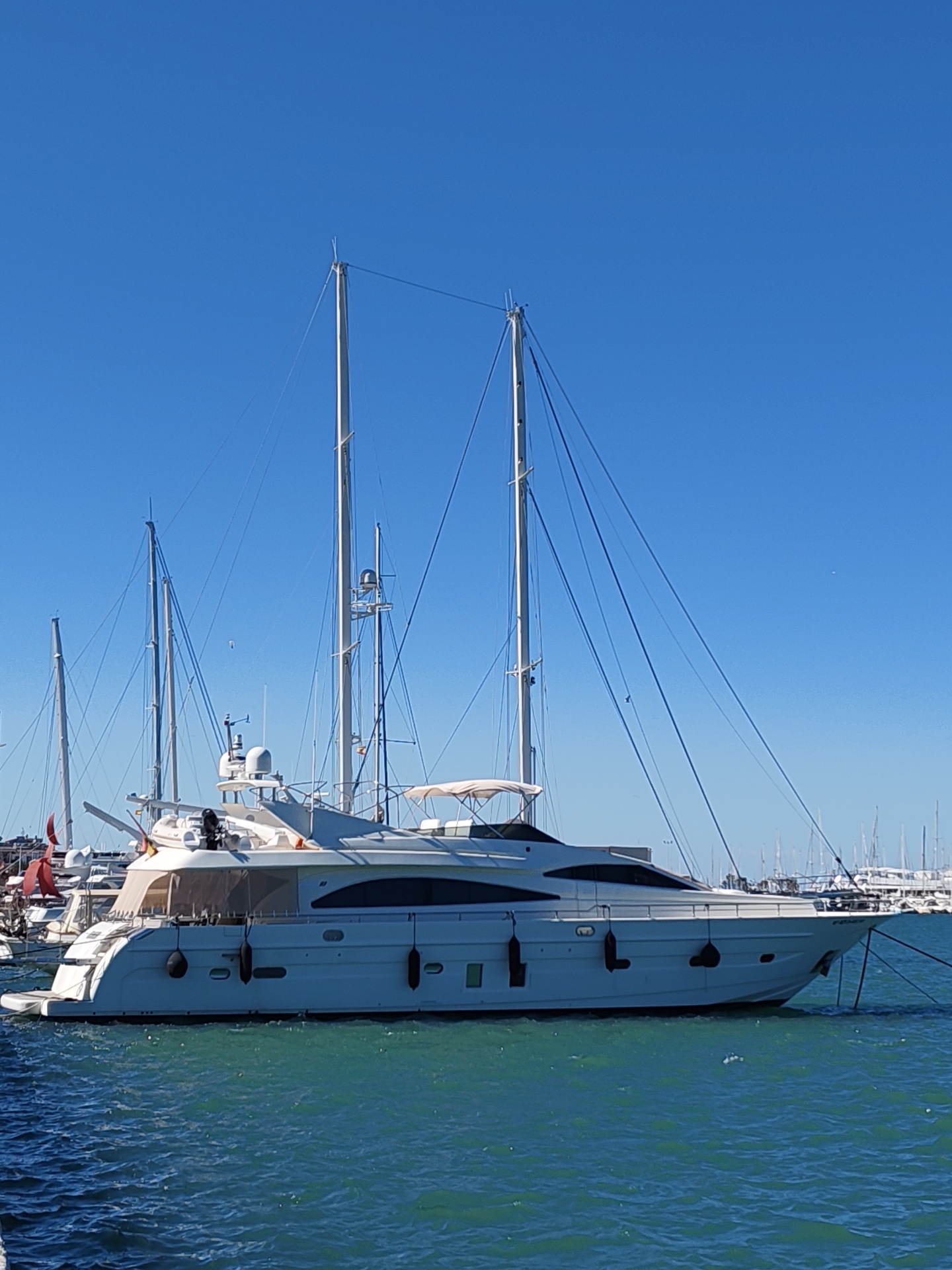 The image depicts a large white yacht moored in the harbor of Dénia, Spain. The yacht has a sleek, modern design with a prominent black stripe along its hull. Three tall masts extend upwards, some with rigging cables. The yacht is docked amongst other vessels, many of which are sailboats with their masts visible in the background. The water is a turquoise color, indicative of a sunny day. The sky is clear and blue. The yacht is secured to the dock with mooring ropes.