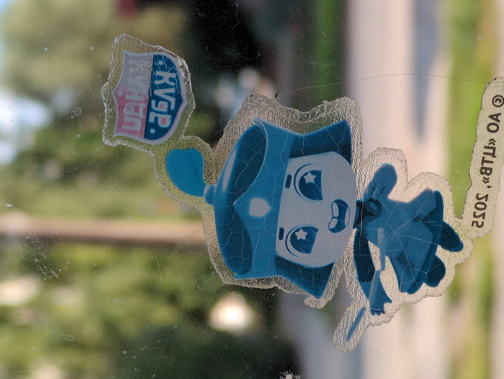 A cartoon sticker, appearing to be on a window, depicts a character wearing a blue turban and holding a sword. The sticker also features a crest with the letters "KVGP" and "UIGHAN" visible. Below the character, a curved text reads "2505, «ВТЦ» ОА ©". The background is blurred, showing greenery and possibly a building, consistent with an outdoor, urban environment.