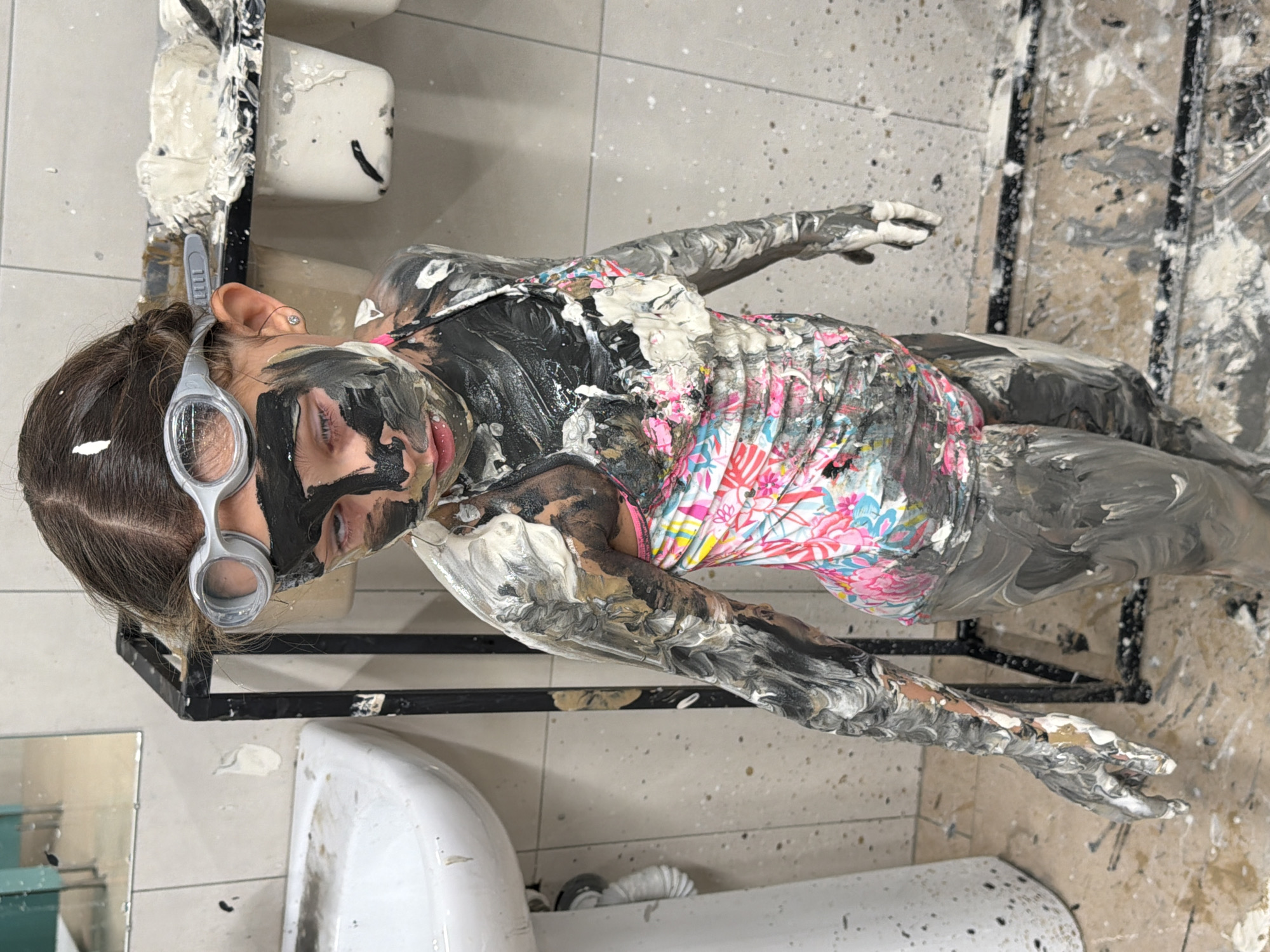 A child covered in paint lies on a tile floor in a bathroom. The child is wearing goggles and a brightly colored floral print shirt. Black, white, and gray paint covers the child's face, arms, legs, and torso. Paint is splattered on the floor and fixtures. A black metal rack is positioned near the child's head. Two white sinks and a mirror are visible in the background. The location in Krasnoyarsk, Russia is not discernible from the image.