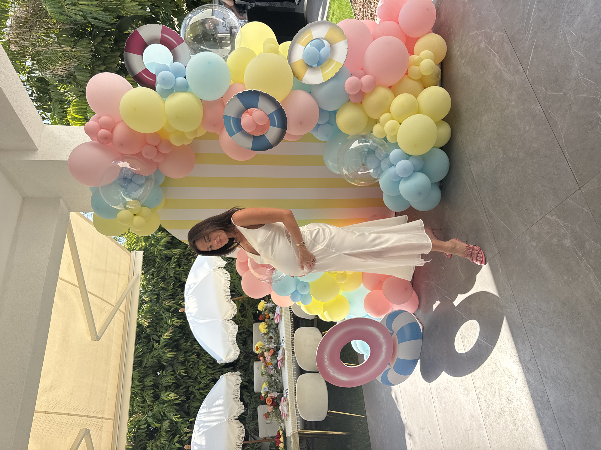 A pregnant woman stands outdoors in front of a balloon arch. She is wearing a long, white, satin dress and pink heeled sandals. Her hands are placed on her pregnant belly. The balloon arch is comprised of pastel-colored balloons in shades of pink, yellow, and blue, and features inflatable life preserver decorations. Behind the woman and the balloon arch is a table set for a meal, with white tablecloths, floral arrangements, and white umbrellas providing shade. The ground is paved with grey tiles. The scene appears to be a celebration, possibly a baby shower, taking place in Agios Tychona Community, Cyprus.