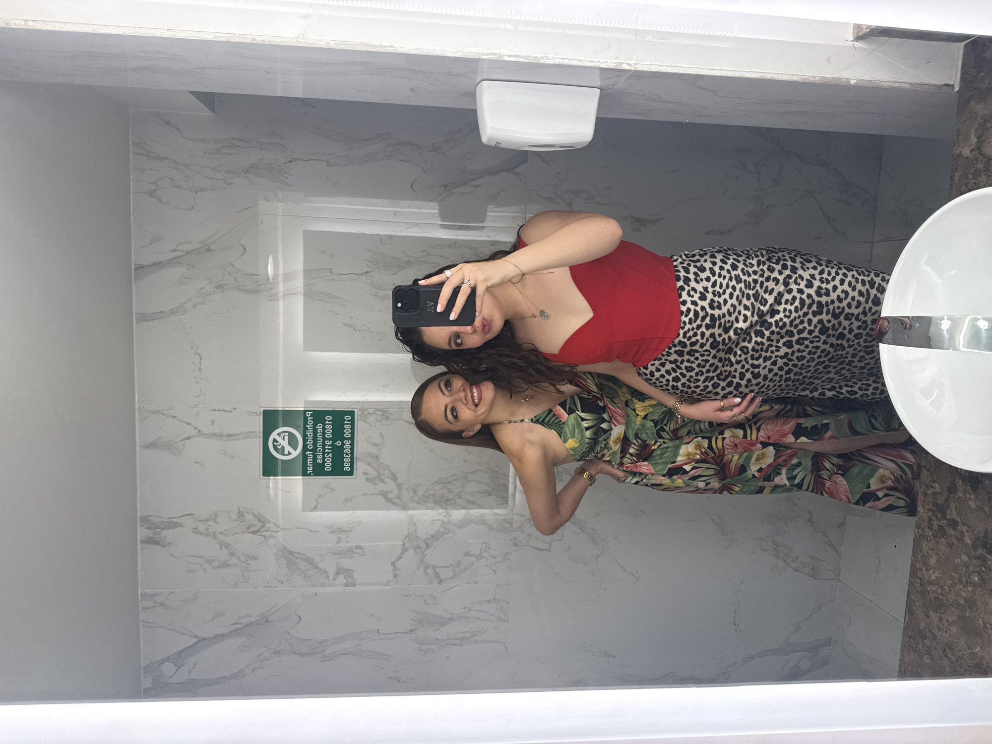 Two women are taking a selfie in a bathroom. The woman taking the photo is wearing a red top and a leopard print skirt. The other woman is wearing a floral dress. The bathroom has marble walls and a white sink. There is a sign on the wall that says "Prohibido Fumar" (No Smoking) and "01800 911 2000" in Spanish. The location may be in Mexico City, Mexico, based on the sign being in Spanish.