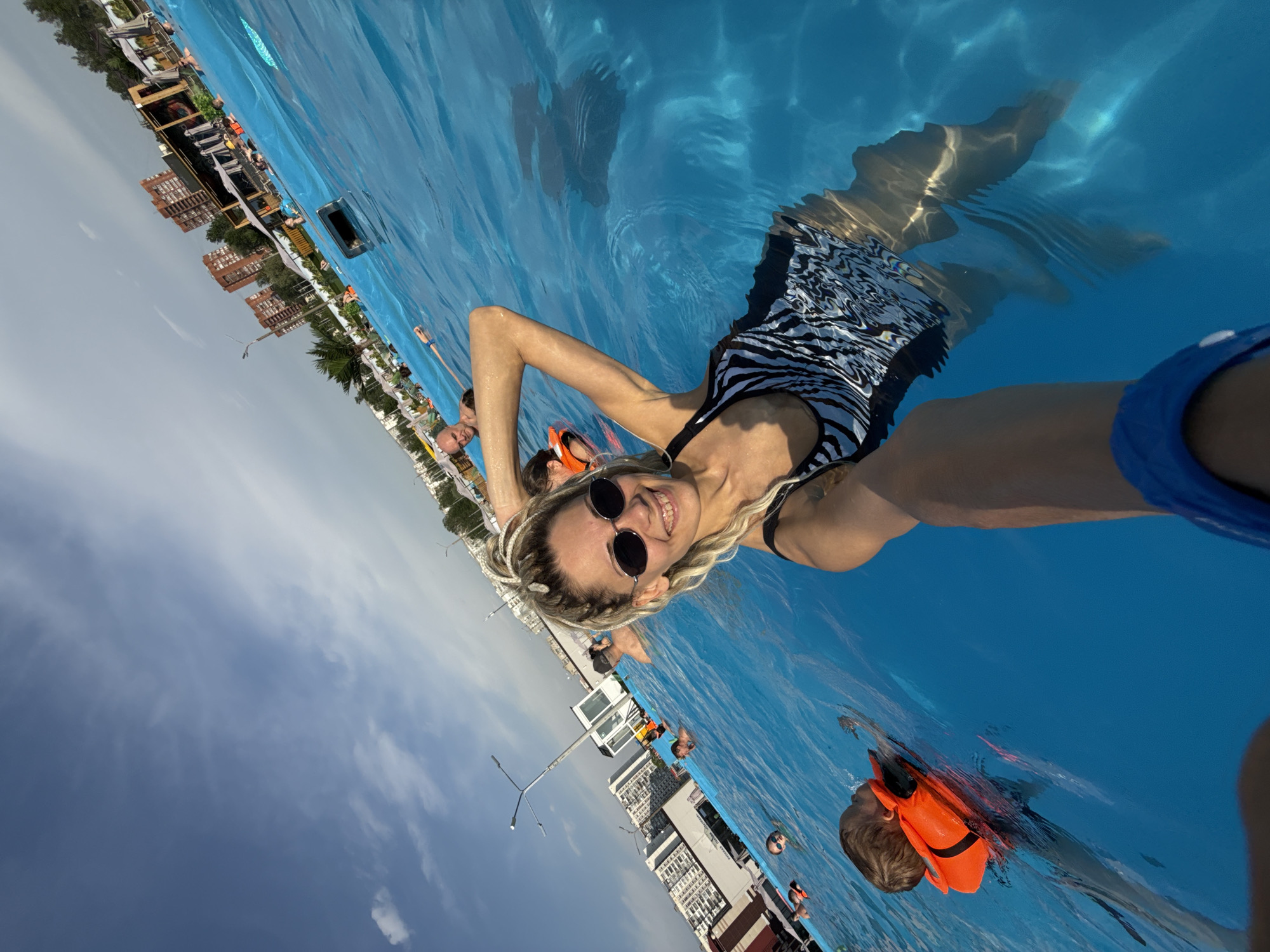 A woman wearing a zebra-striped bathing suit and sunglasses is in a pool of blue water, taking a selfie. The cityscape of Rusanovo, Russia is visible in the background, including buildings and a partly cloudy sky. Other people are in the pool, including a child wearing an orange life vest.