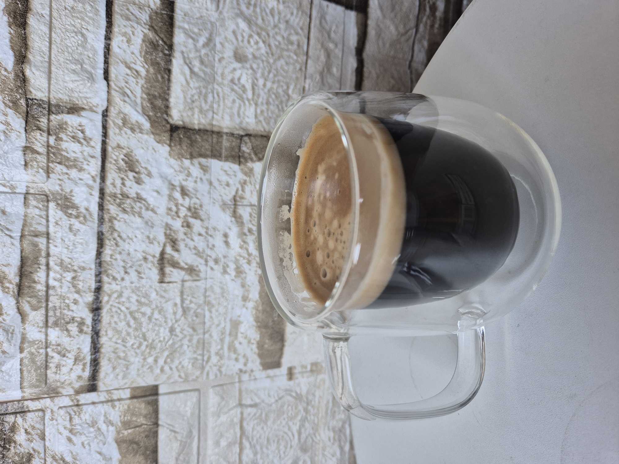 A clear glass mug filled with dark coffee sits on a white surface. The mug has a clear handle and a double-walled design. The coffee has a foamy, light brown crema on top. In the background, a textured wall with a faux brick pattern is visible. The scene is set in an indoor environment, likely a home or cafe. No people are present in the image.