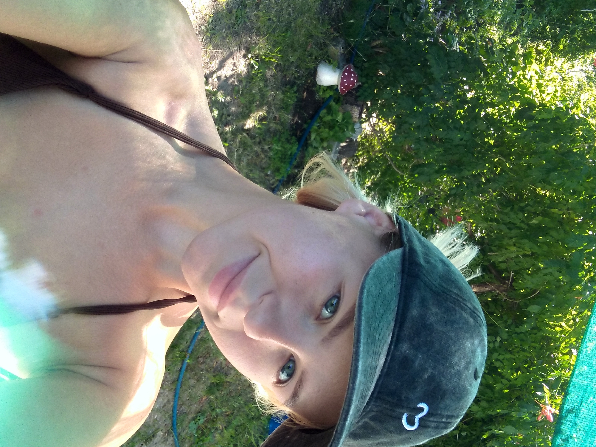 A young woman is taking a selfie outdoors in what appears to be a garden or yard. She is wearing a dark green baseball cap and a brown bikini top. Her blonde hair is visible under the cap. She has light skin and blue eyes. The background shows lush green foliage, grass, and a small decorative mushroom. Sunlight filters through the trees. The overall setting is bright and natural. The specific location is in the city of Radisne, Ukraine.
