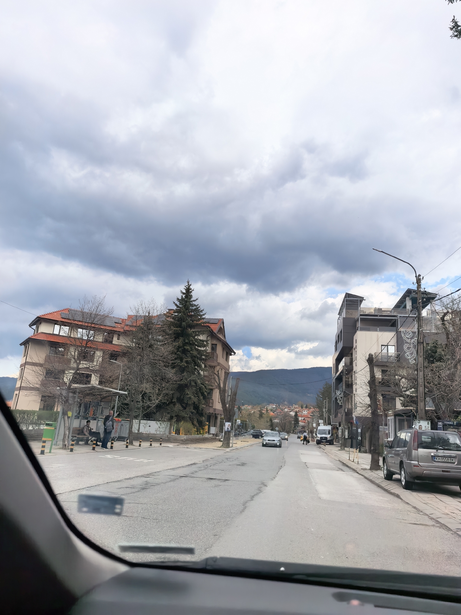 The image depicts a street scene in a city. The view is from inside a car, looking out through the windshield. The street is paved and has buildings on either side. A large multi-story building is on the left with an orange roof and light-colored exterior. A bus stop sits at the corner with people waiting under it. A green evergreen tree is to the right. On the right side of the street, there is a modern building with gray and tan colors. A gray SUV is parked along the curb. Other cars and pedestrians are visible further down the street. Mountains are visible in the distance beyond the buildings. The sky is overcast with gray clouds. The location is Unknown, Unknown.