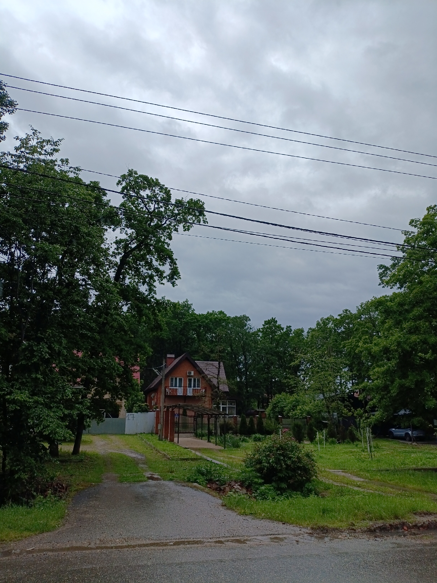 The photograph depicts a residential scene in Goryachiy Klyuch, Russia, under an overcast sky. A two-story brick house with a dark red roof is partially obscured by lush green trees. The house features two white-framed windows with small balconies on the second floor and a covered porch area. Electrical wires are visible stretching across the sky. A dirt and grass driveway leads from the street to the house. The foreground shows a wet asphalt road with grassy edges. A small bush and other greenery are present on the side of the driveway. A fence can be seen in the background. A car is parked on the grass to the right of the frame.
