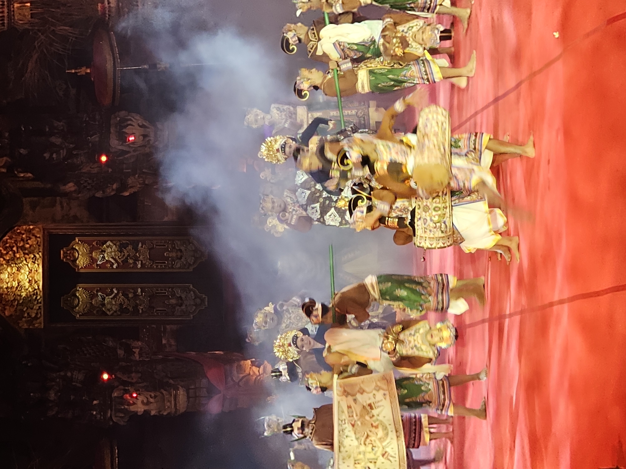 The image depicts a cultural performance taking place on a vibrant red stage, likely in Denpasar, Indonesia, given the context of traditional Balinese dance. A group of performers, both male and female, are dressed in elaborate, colorful costumes adorned with intricate patterns and golden headdresses. They are engaged in a dance, with some holding long green poles and others carrying large, patterned fabric scrolls. Smoke or fog drifts across the stage, adding a dramatic atmosphere to the scene. In the background, a detailed, ornate golden structure with carved figures and two large doors is visible, suggesting a temple or a significant cultural venue. The lighting is theatrical, with red hues and spotlights illuminating the performers and the stage.