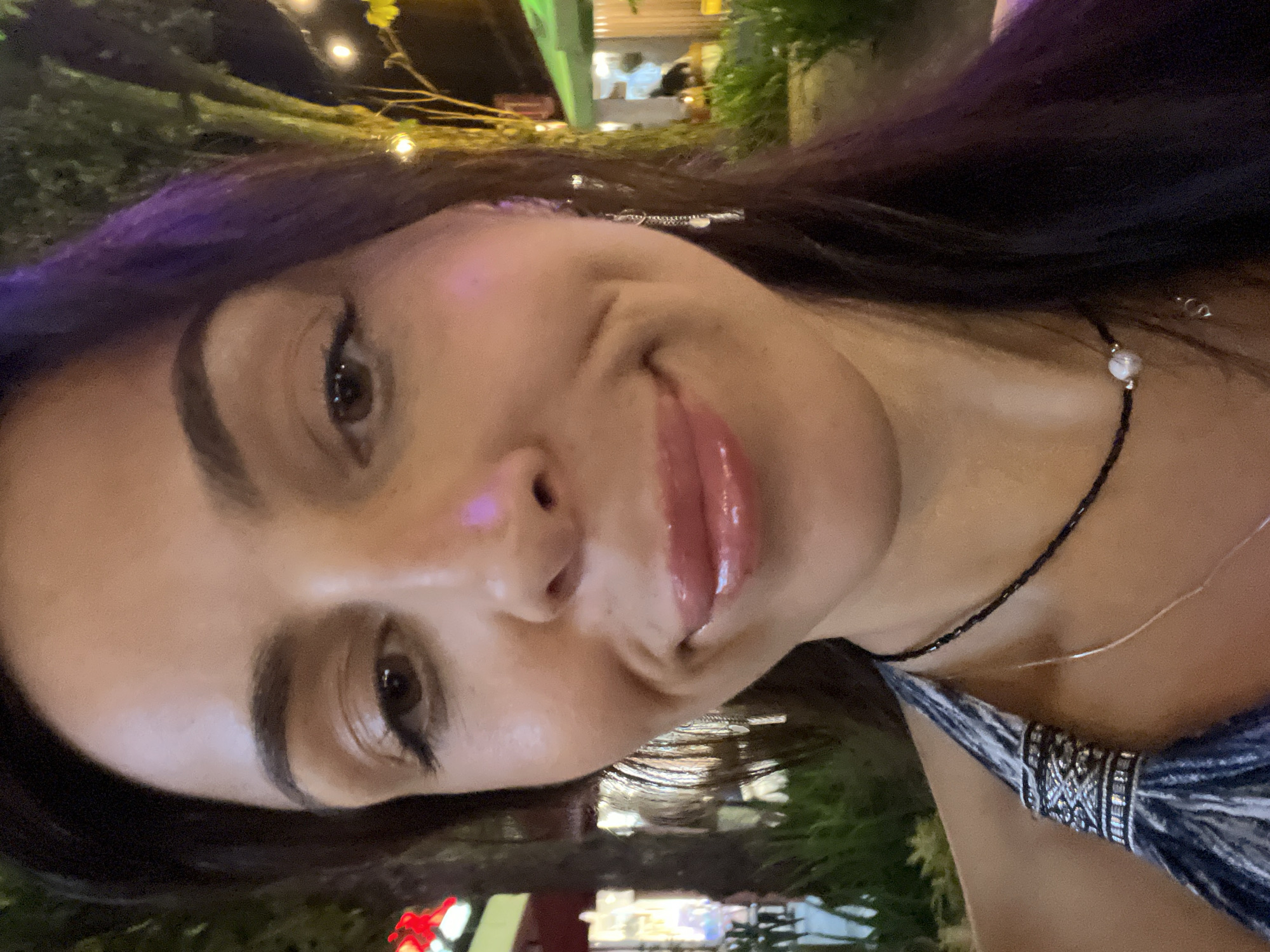 A selfie-style photograph captures a woman with long, dark hair, featuring purple highlights at the top. Her skin tone appears light to medium, and she has dark eyebrows and brown eyes. She is wearing a black beaded necklace with a single white bead. Her lips are glossy and appear to be slightly pursed. A patterned garment is visible on her right shoulder.
The background is blurred, suggesting an outdoor setting at night. There are hints of greenery and indistinct light sources that could be urban lighting. The location is stated to be Unknown, Indonesia, but there are no distinguishing landmarks or features in the image to confirm or specify a more precise location.