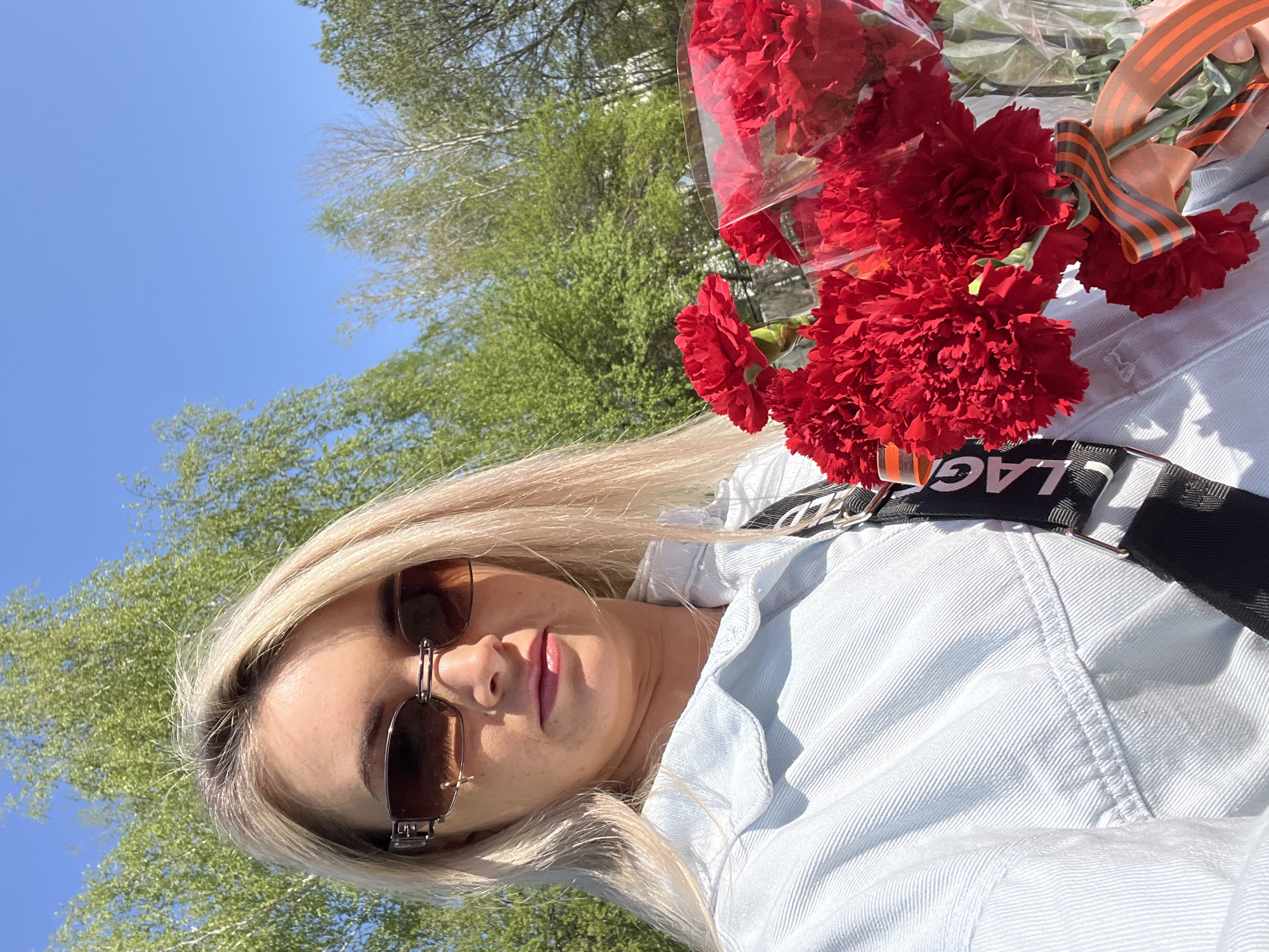 A young woman is holding a bouquet of red carnations tied with a St. George ribbon. She is wearing sunglasses and a light blue jacket. The scene is outdoors with trees and a blue sky visible in the background. The photo was taken in Chaikovsky, Russia.