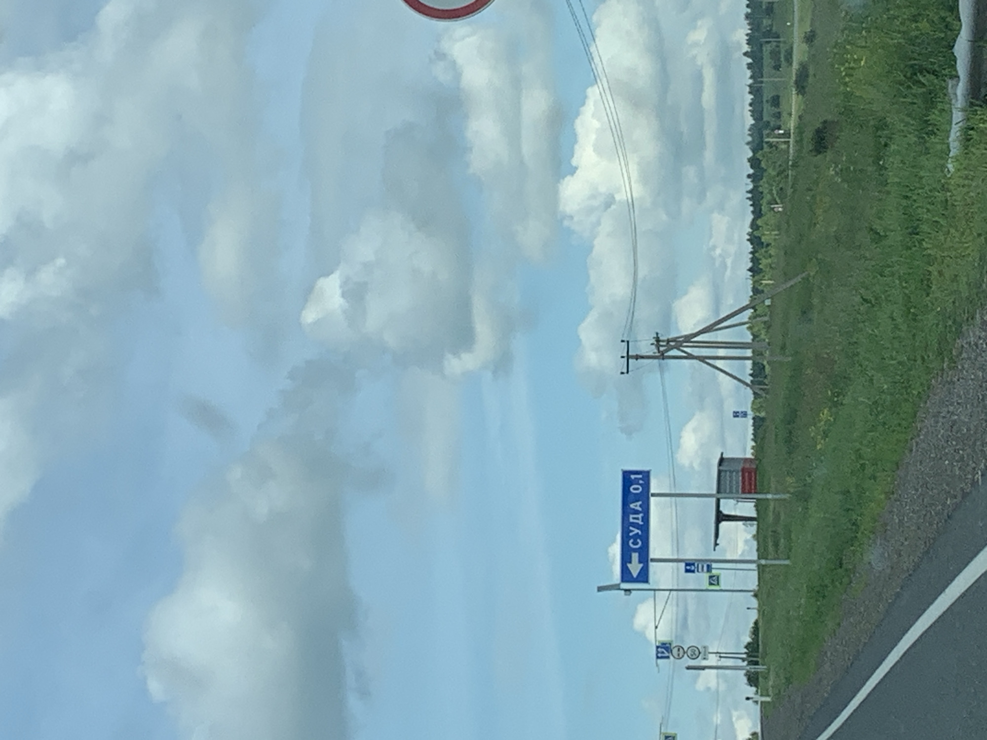 A road sign in the city of Суда, Russia, indicates a distance of 0.1 kilometers in a direction to the left. The sign is blue with white text and an arrow pointing left. In the background, there is a green grassy field with trees and a cloudy sky. Several other signs, including a parking sign and a speed limit sign, are visible near the main road sign. There are also power lines and a small bus stop structure in the distance. The perspective suggests the photo was taken from a vehicle driving on the road.